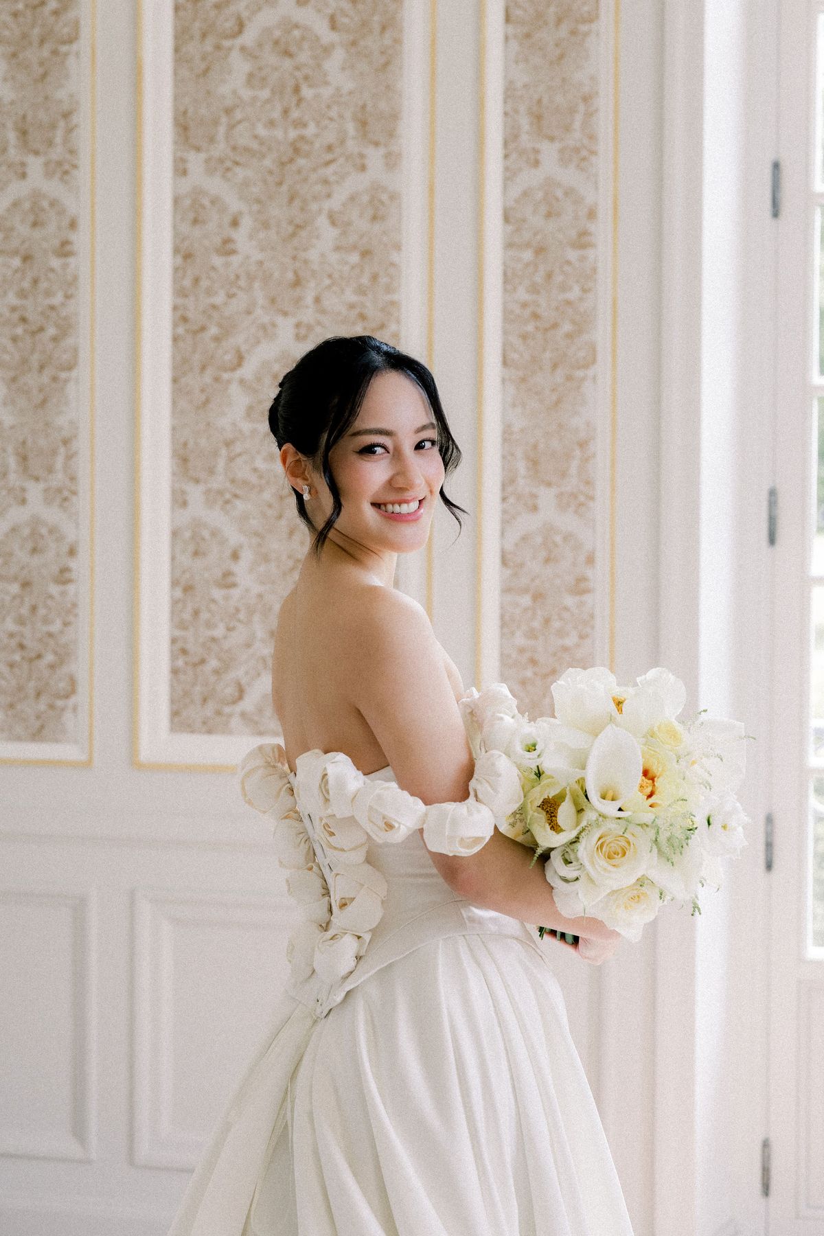 The bride smiles over her shoulder holding a bouquet of white calla lilies and roses