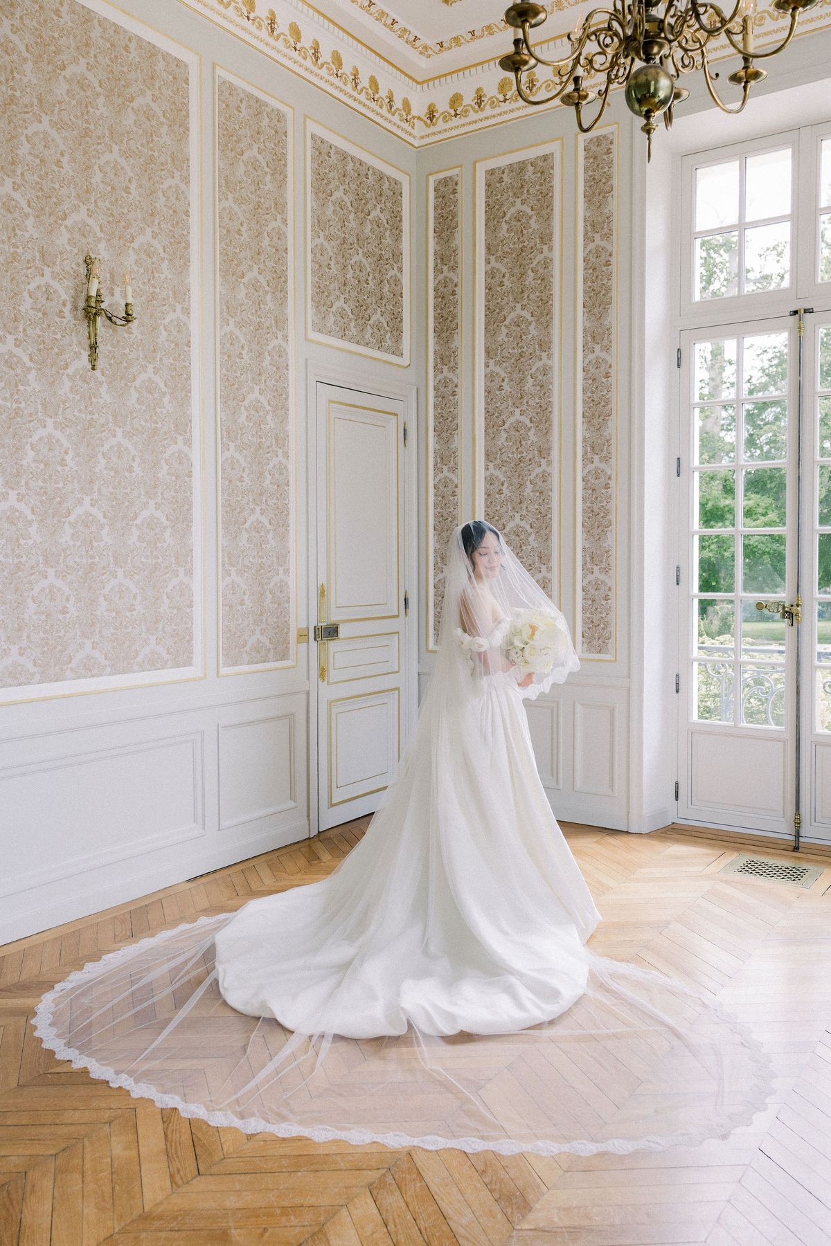 The bride in her veil stands in a damask-panelled salon with the long veil circled around her