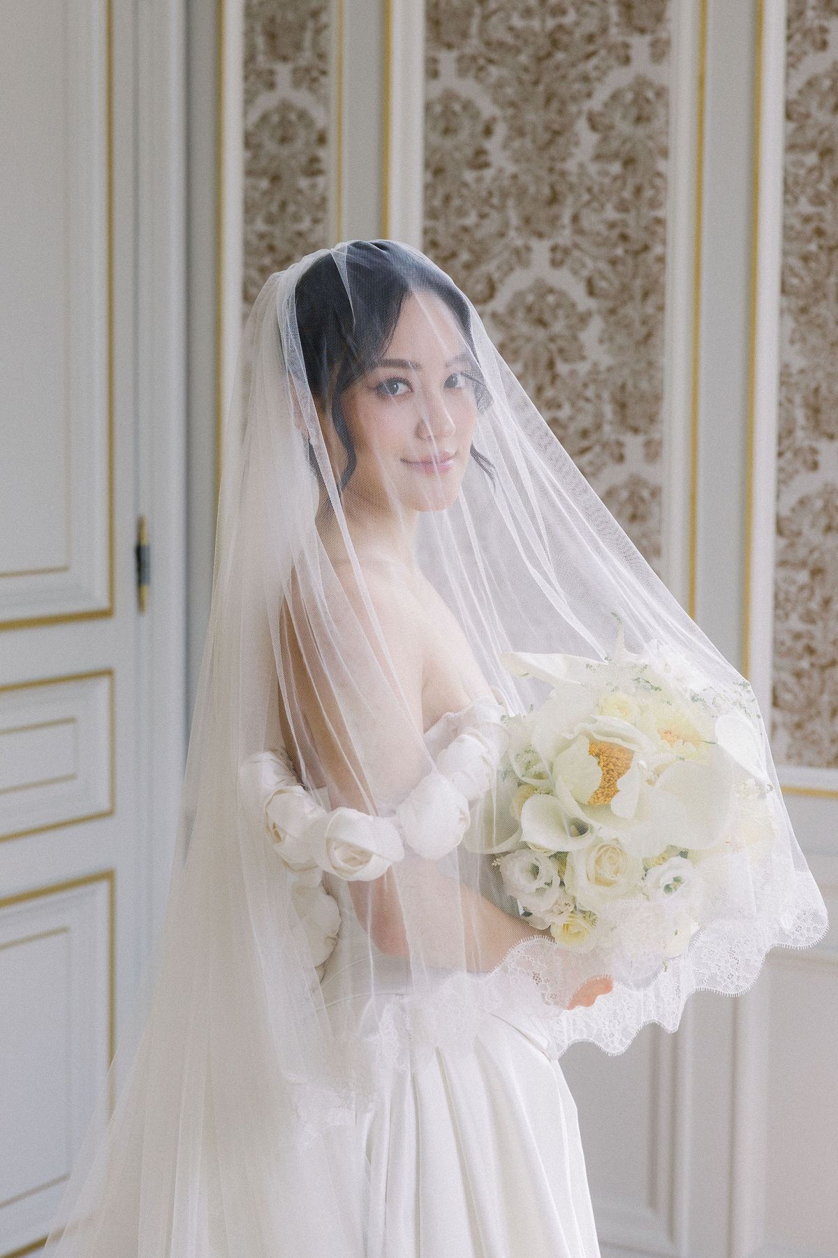 The bride looks out from under her veil holding a white bouquet
