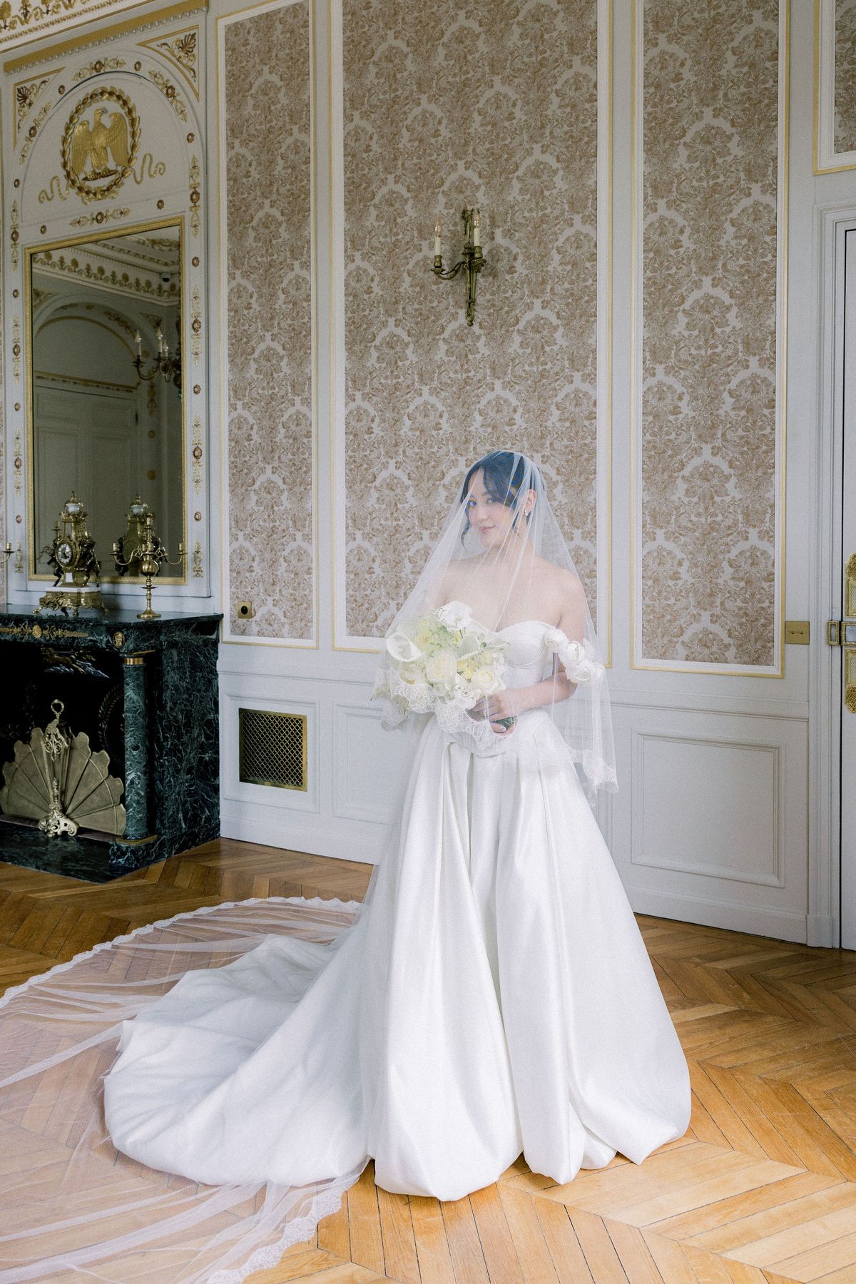 The bride in her veil beside a green marble fireplace in a gilded chateau salon