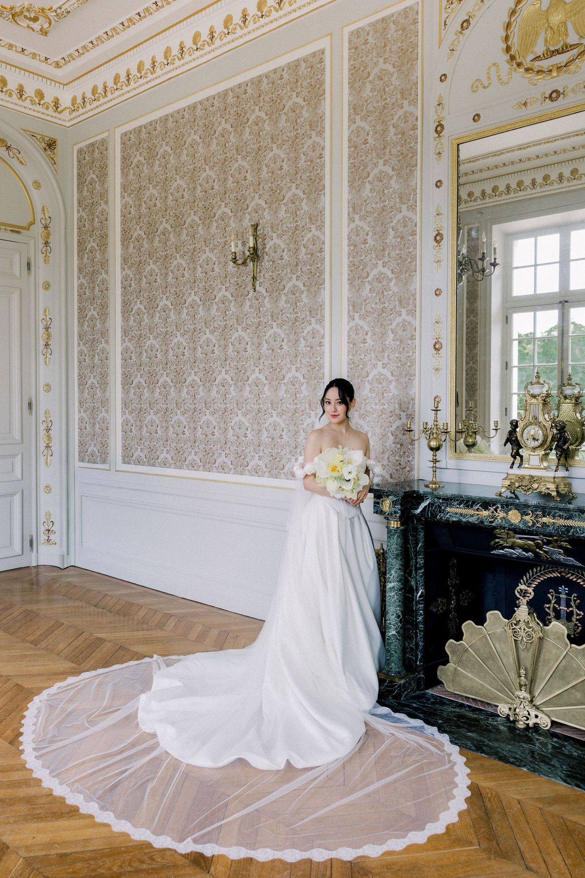 The bride stands by a green marble fireplace with her lace-edged veil laid in a circle on the floor
