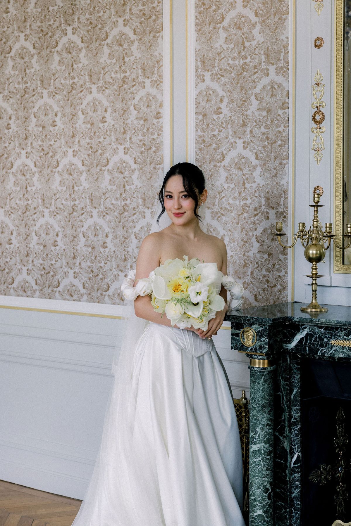 The bride holds her bouquet beside a gilt clock on a green marble mantelpiece