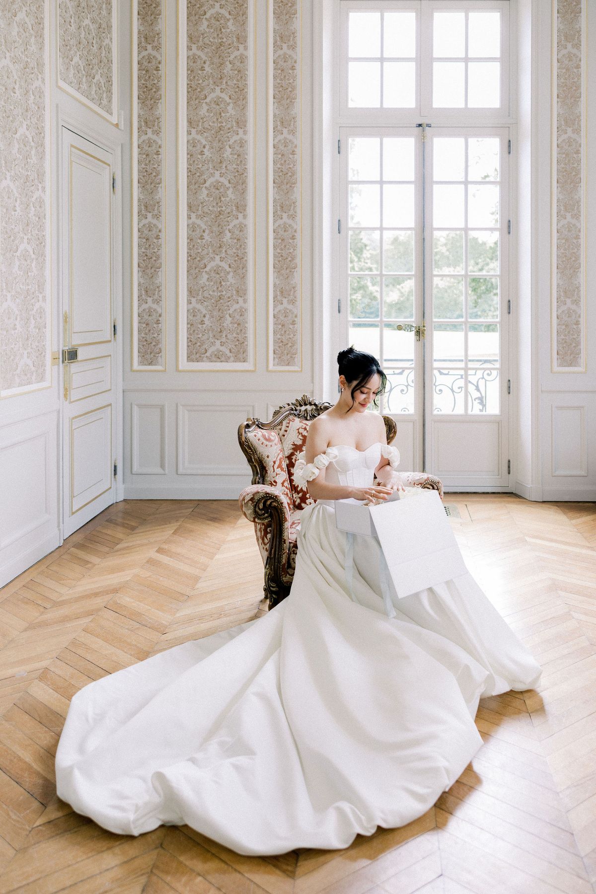 The bride sits on a carved armchair in a bright salon with an open gift box at her feet
