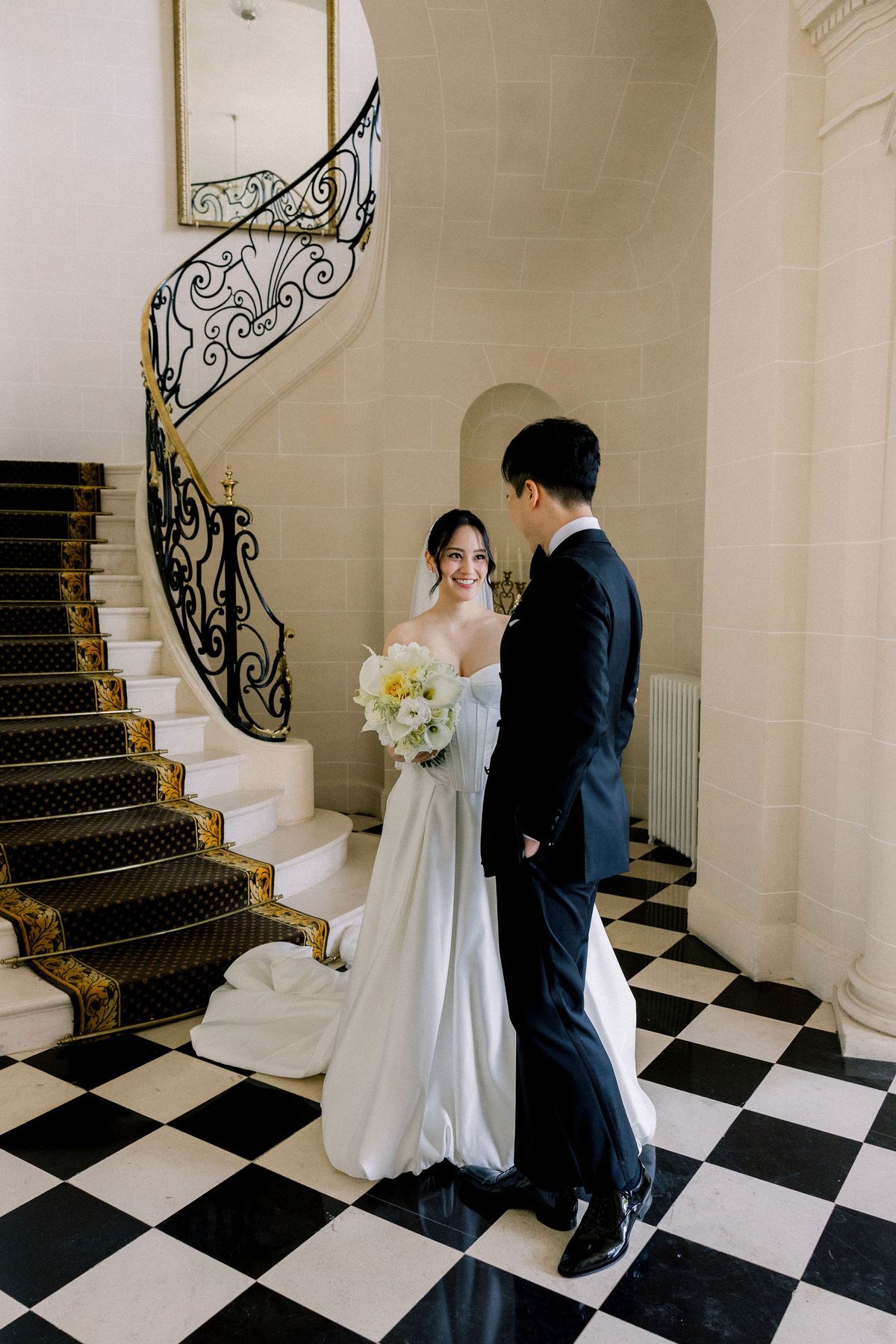 The couple face each other on the black and white chequerboard floor at the foot of the staircase