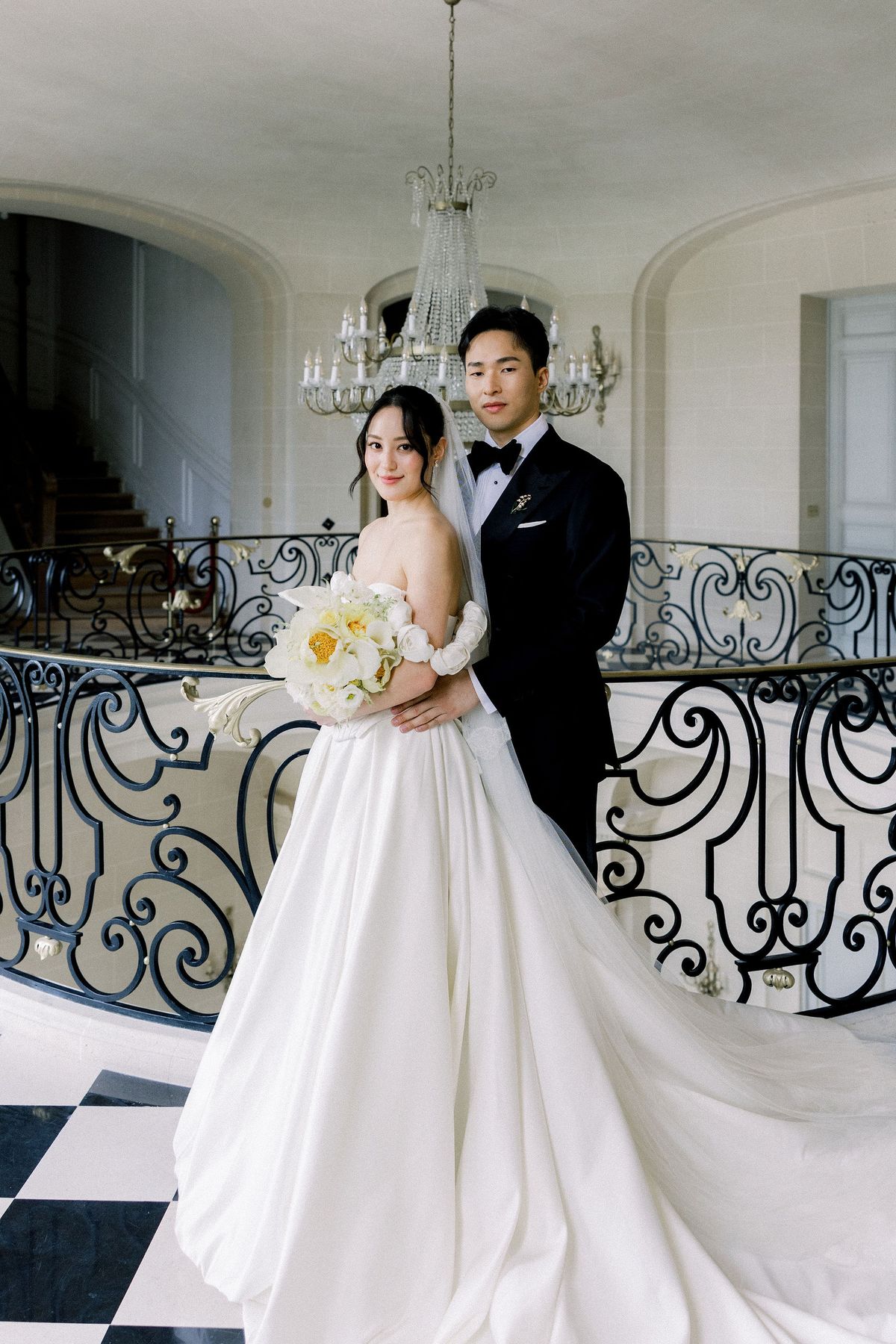 The couple pose at the iron balustrade of the upper landing beneath a crystal chandelier