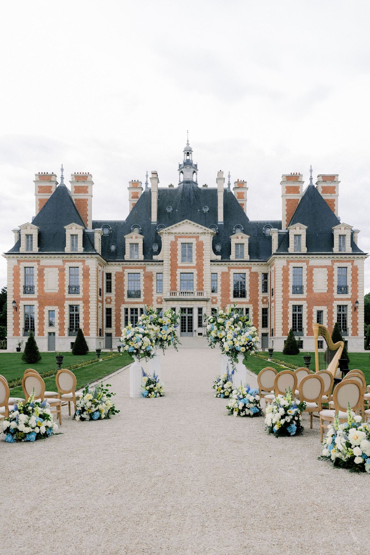 The ceremony aisle of gravel, flanked by wooden chairs and blue and white flowers, leading to the chateau facade