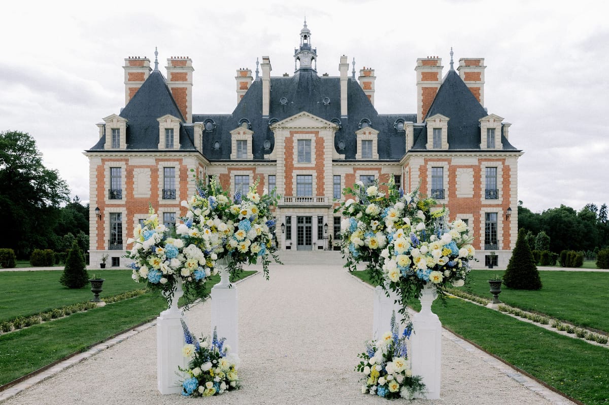 Two tall white plinths of white, yellow and blue flowers frame the aisle in front of the chateau