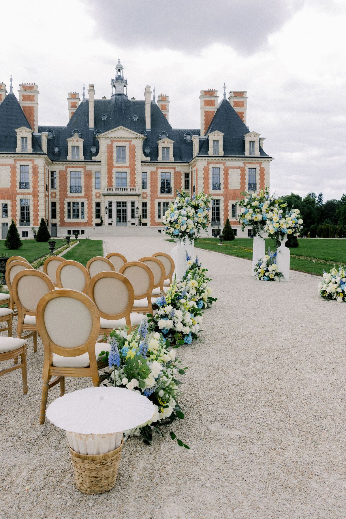 Rows of wooden chairs with upholstered oval backs and aisle flowers before the chateau facade