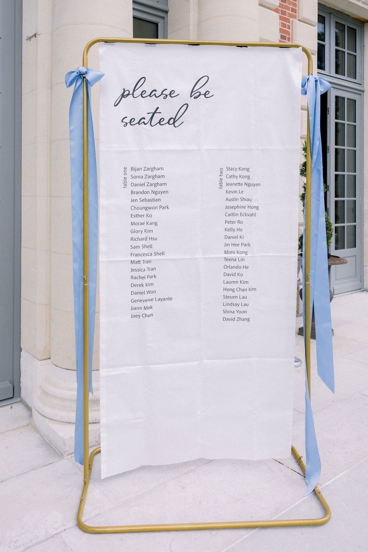 A seating plan on white fabric in a gold frame tied with pale blue ribbons