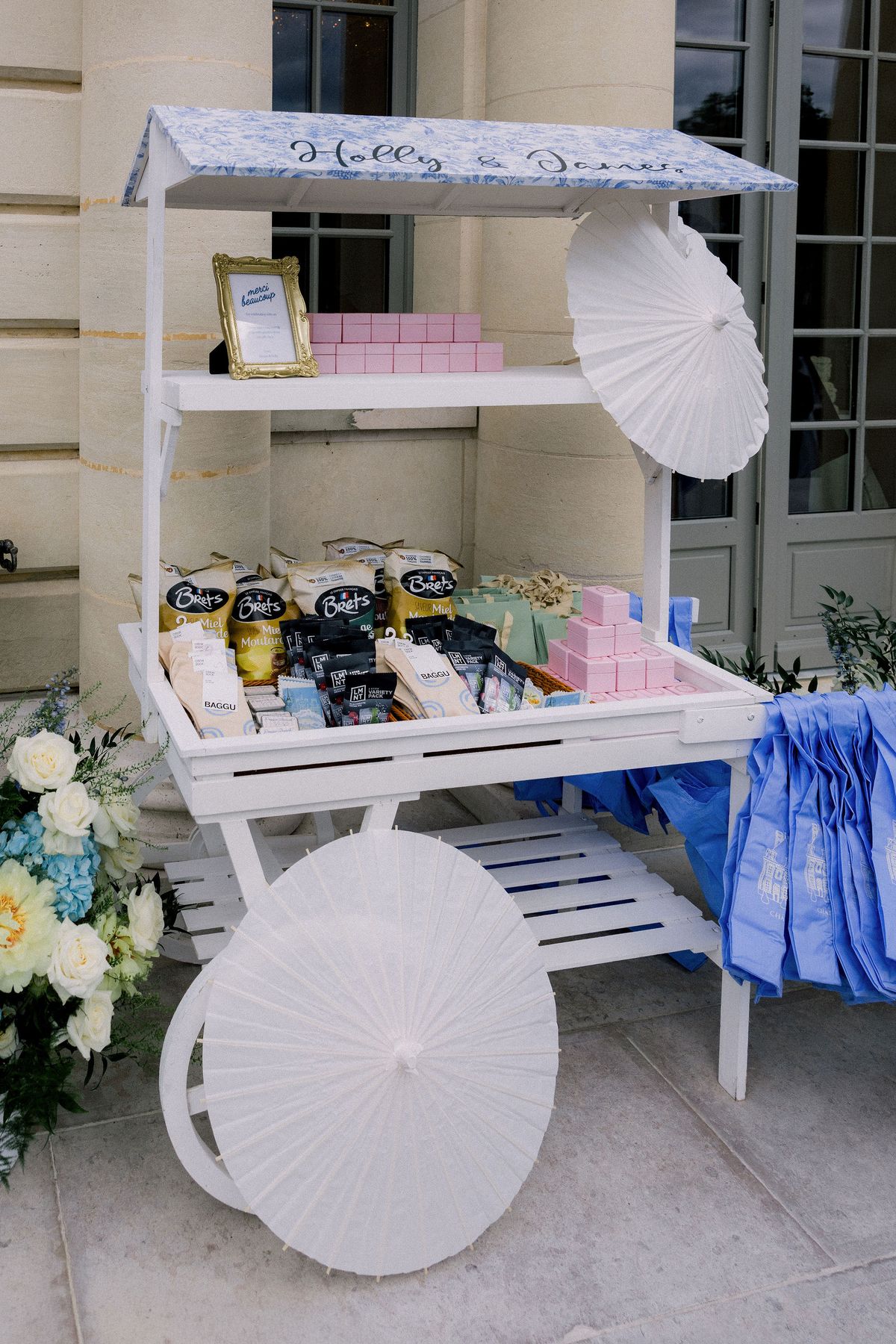 A white wooden favour cart with paper parasols under a blue toile awning outside the chateau