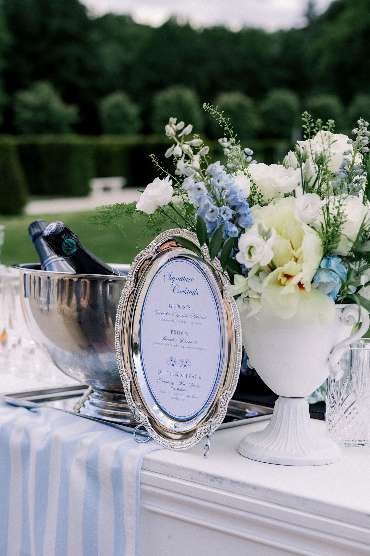 A silver-framed signature cocktail menu beside a champagne bucket and blue and white flowers
