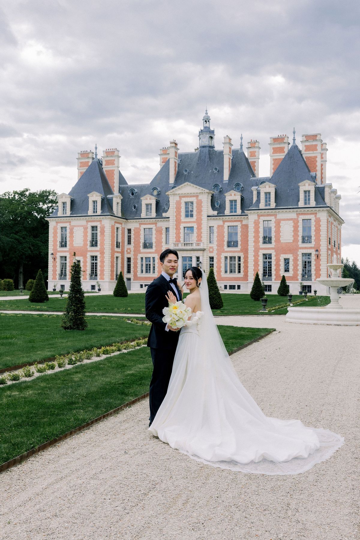 The couple stand on the gravel drive with the chateau and clipped yews behind them