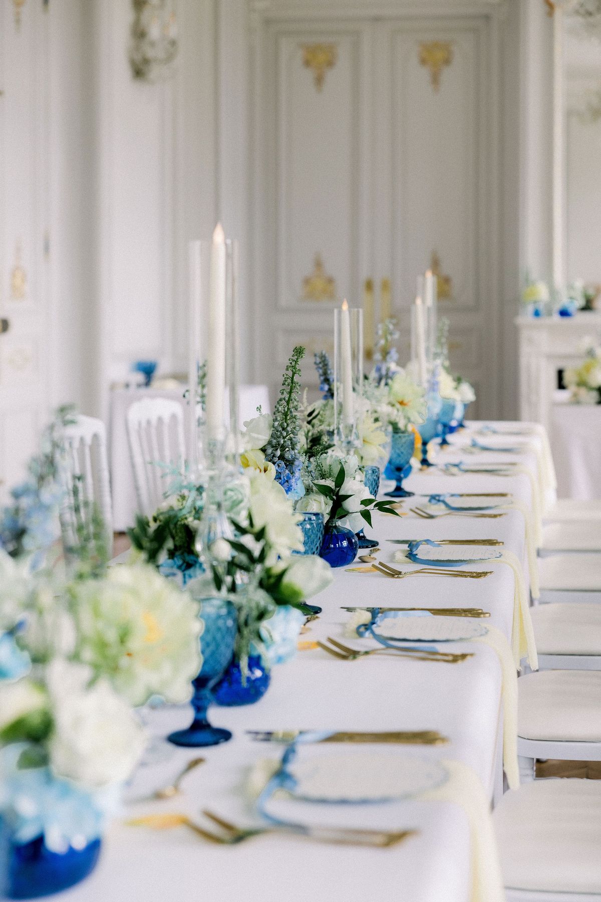 A long dinner table with tall white candles, blue glassware and low flowers in a white and gold salon