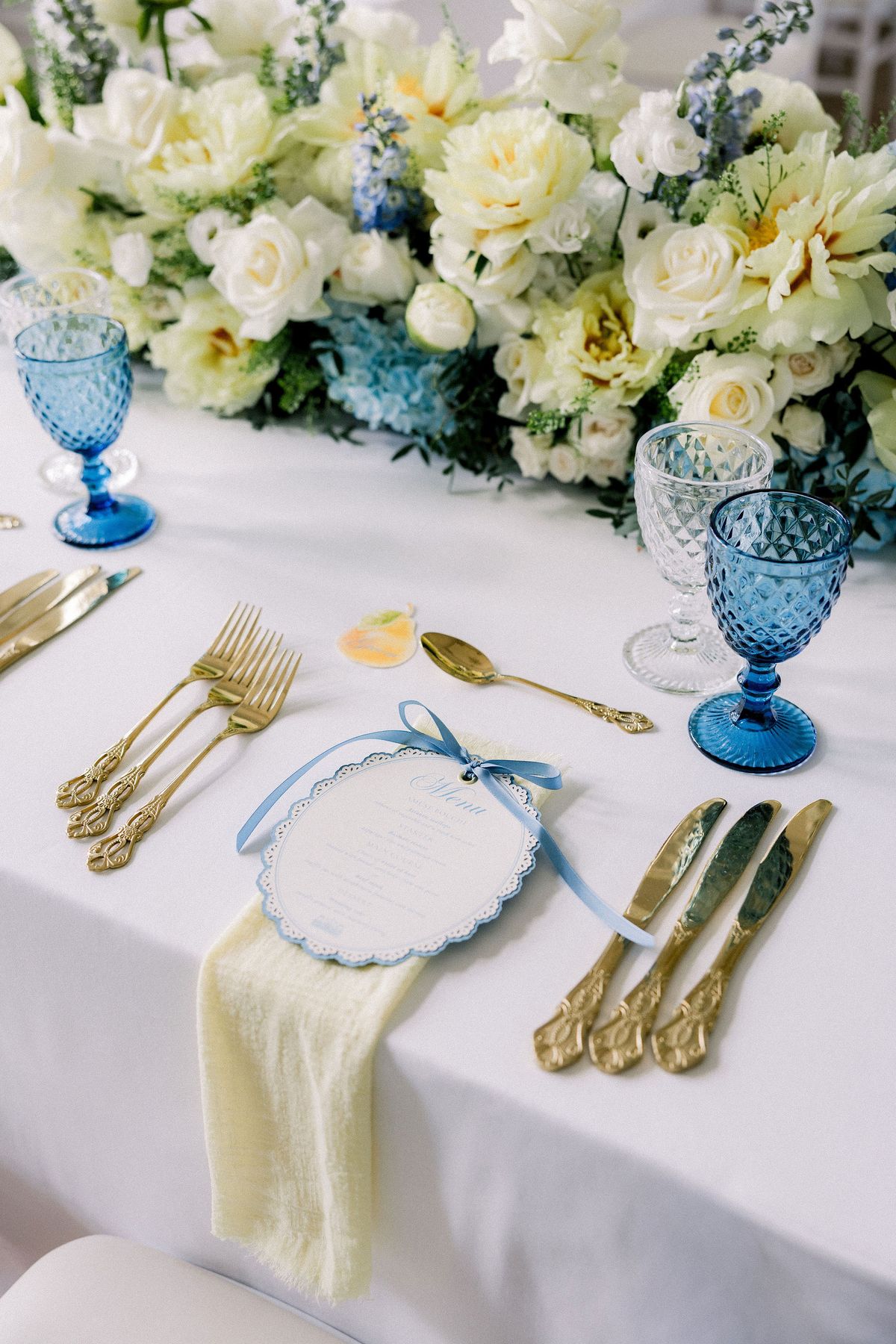 A place setting with a scalloped blue menu, gold cutlery and a pale yellow napkin beneath white flowers