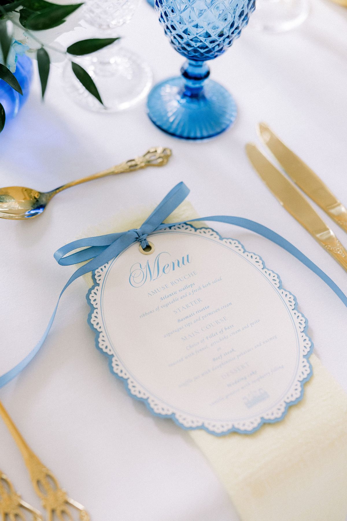 A blue scalloped menu tied with ribbon beside a blue goblet and gold cutlery