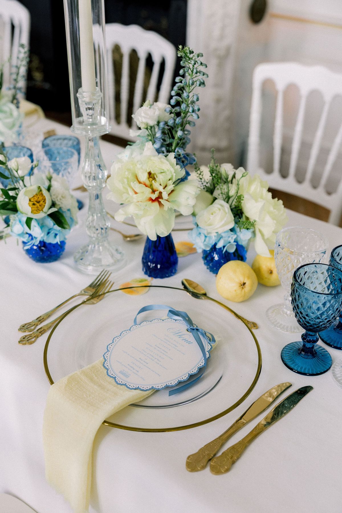 Blue bud vases, a small yellow fruit and a ribboned menu on a gold-rimmed charger plate