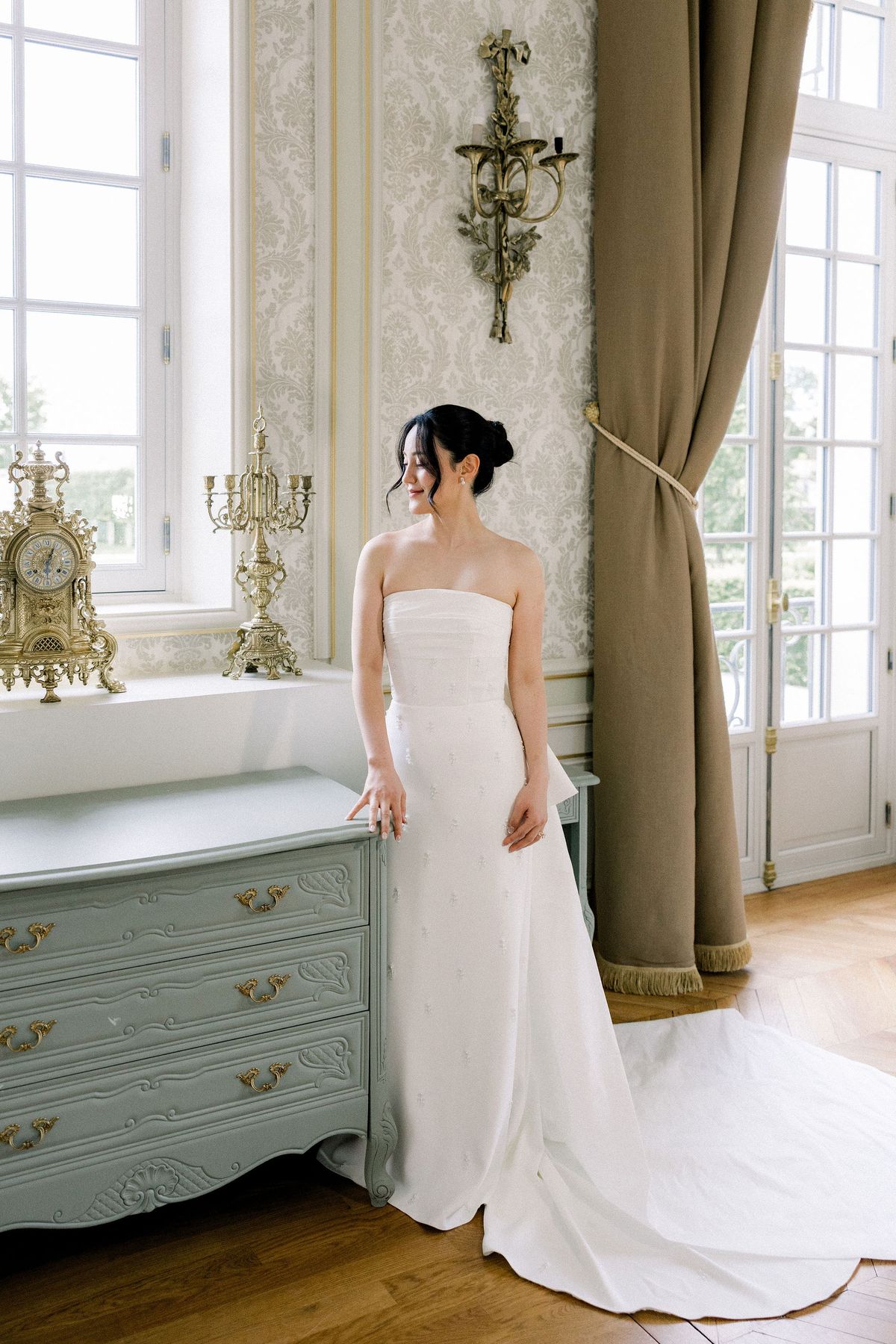 The bride in her fitted evening dress stands at a tall window beside a pale green chest of drawers