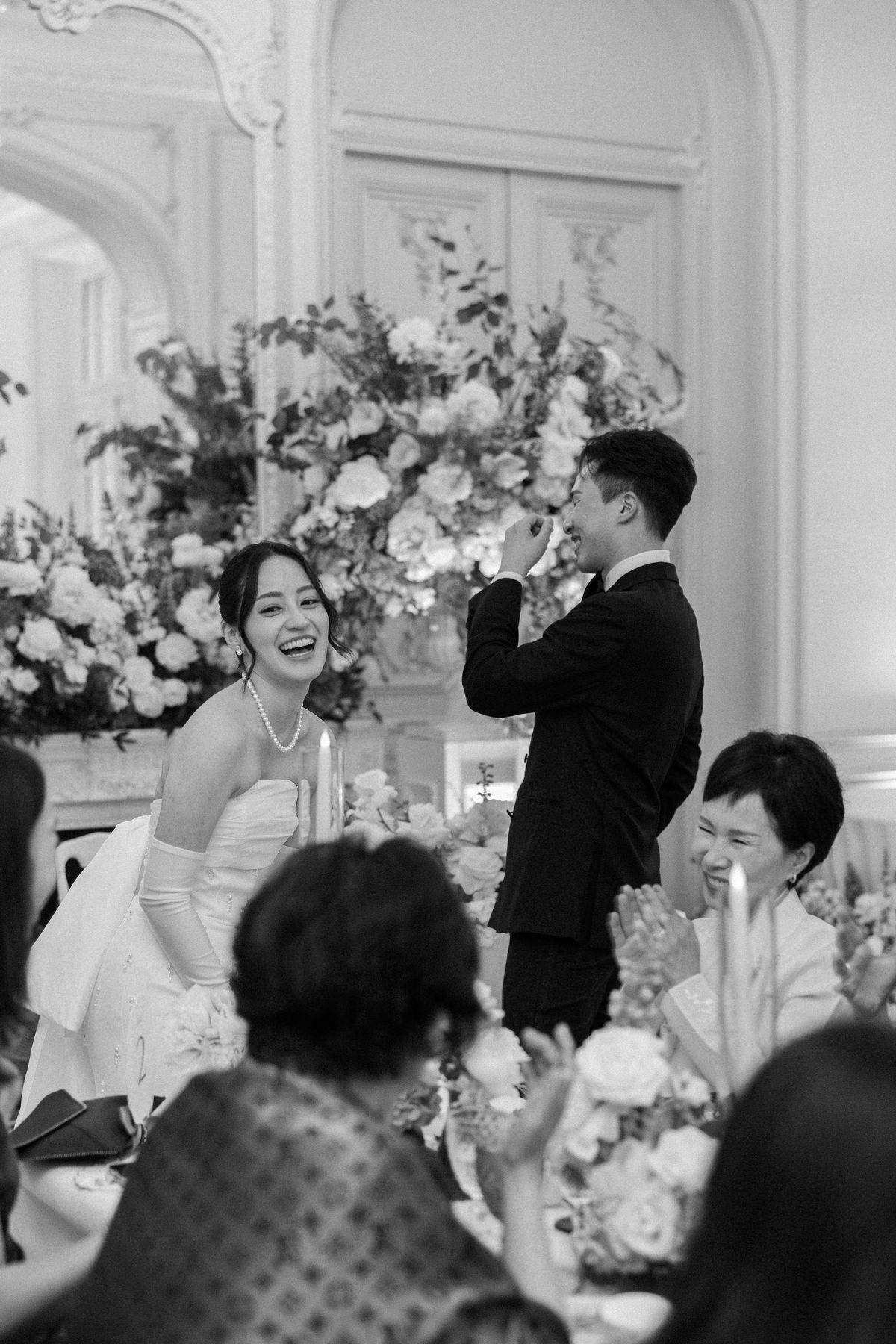 Black and white image of the bride laughing as the groom raises a hand to his face at dinner