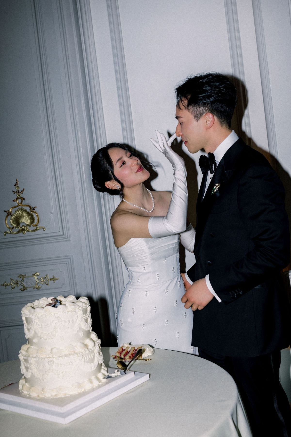 The bride in long white gloves feeds the groom a taste of the two-tier white cake