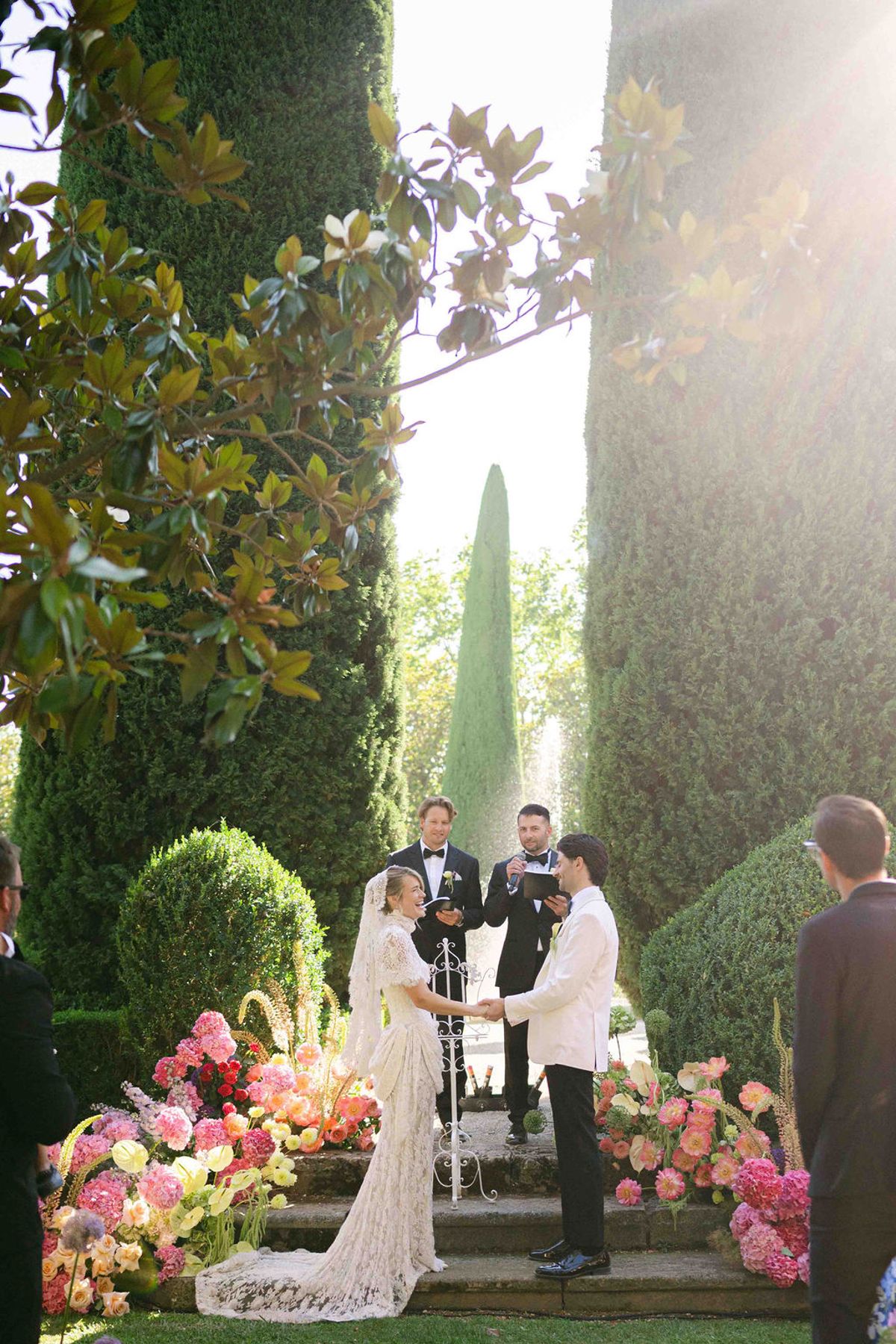 The couple hold hands at a flower-lined altar between tall cypress trees, lit by soft sunlight from behind.