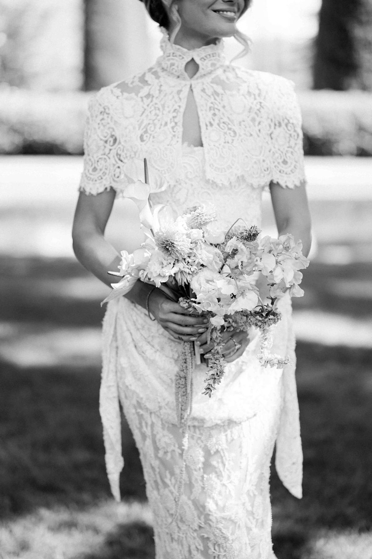 A black and white image of the bride in a lace dress with a high collar, holding a loose bouquet.