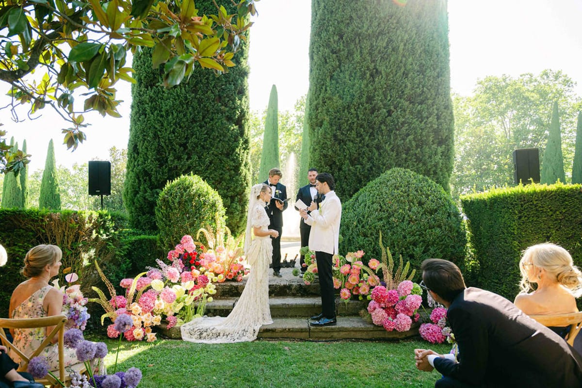 The couple stand before an officiant on garden steps lined with pink flowers while guests sit watching on either side.