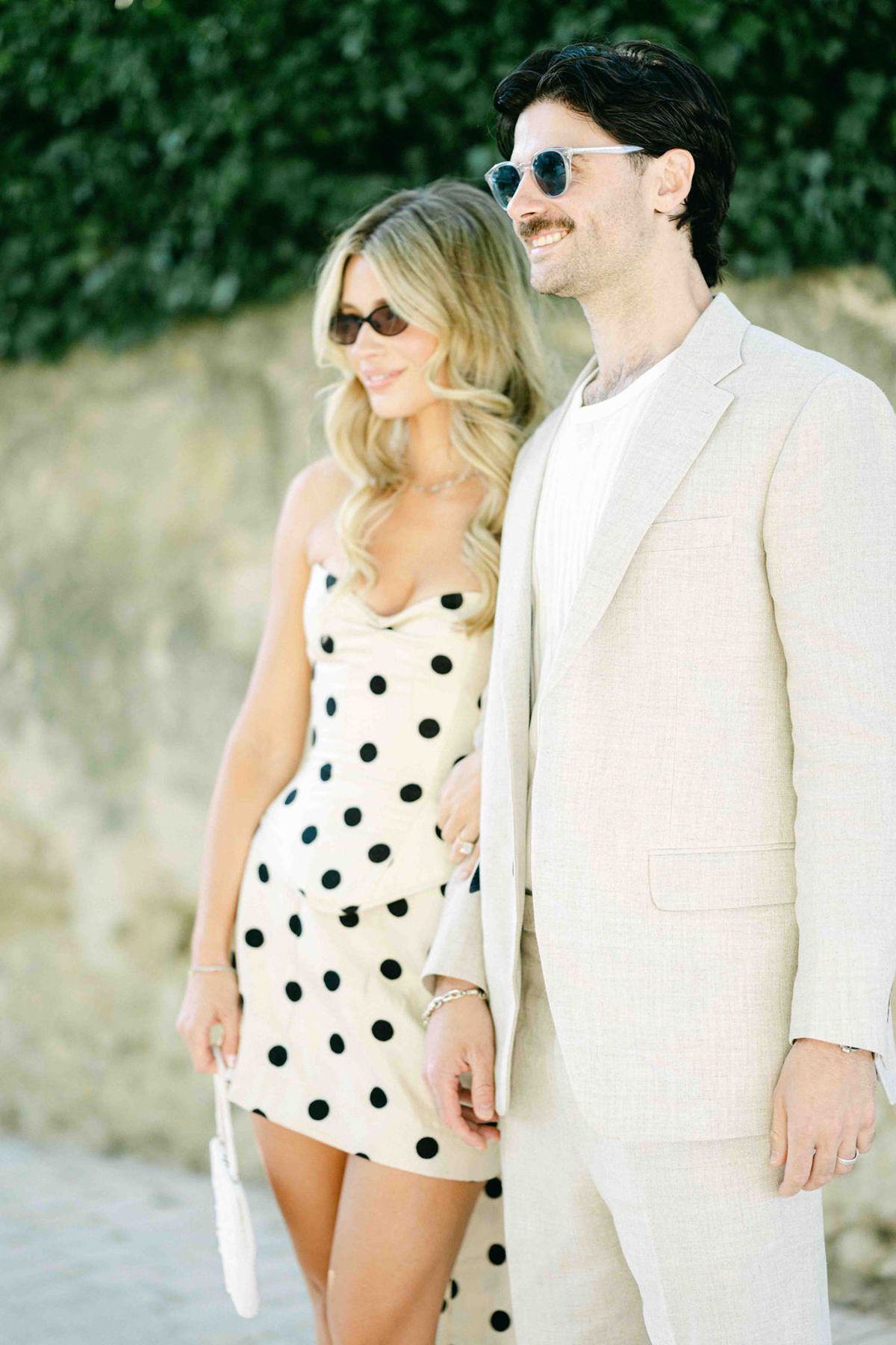 The couple stand close together wearing sunglasses, one in a polka dot dress and the other in a cream suit.