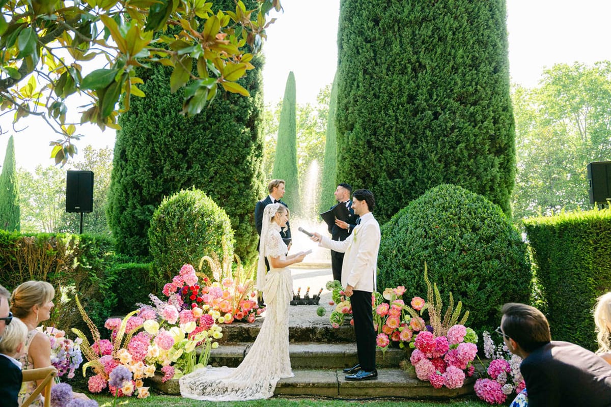 The bride and groom stand before an officiant on garden steps lined with pink flowers, with guests seated either side.