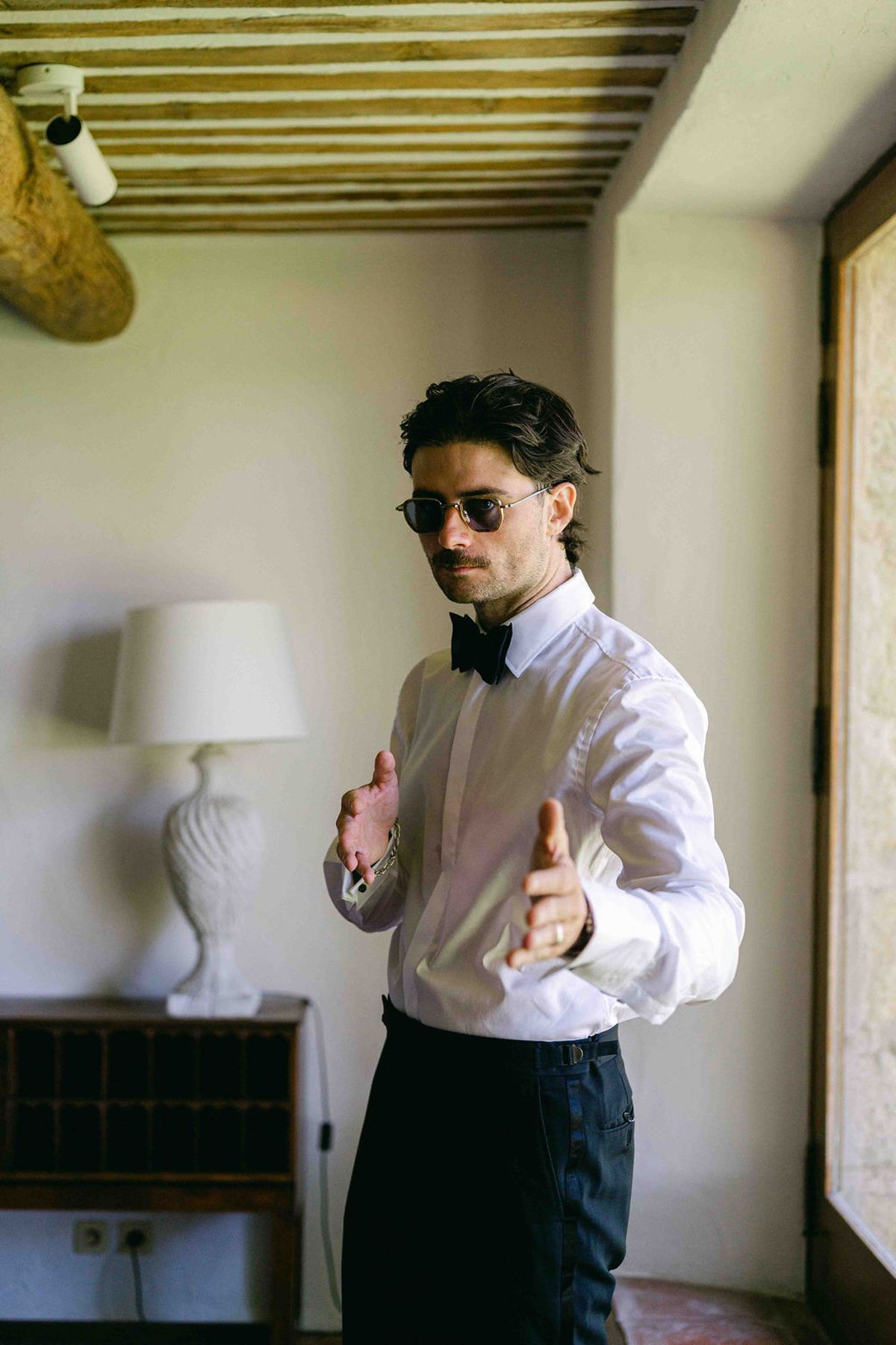 The groom, wearing sunglasses, a white shirt and a black bow tie, gestures with both hands in a bright room with a beamed ceiling.