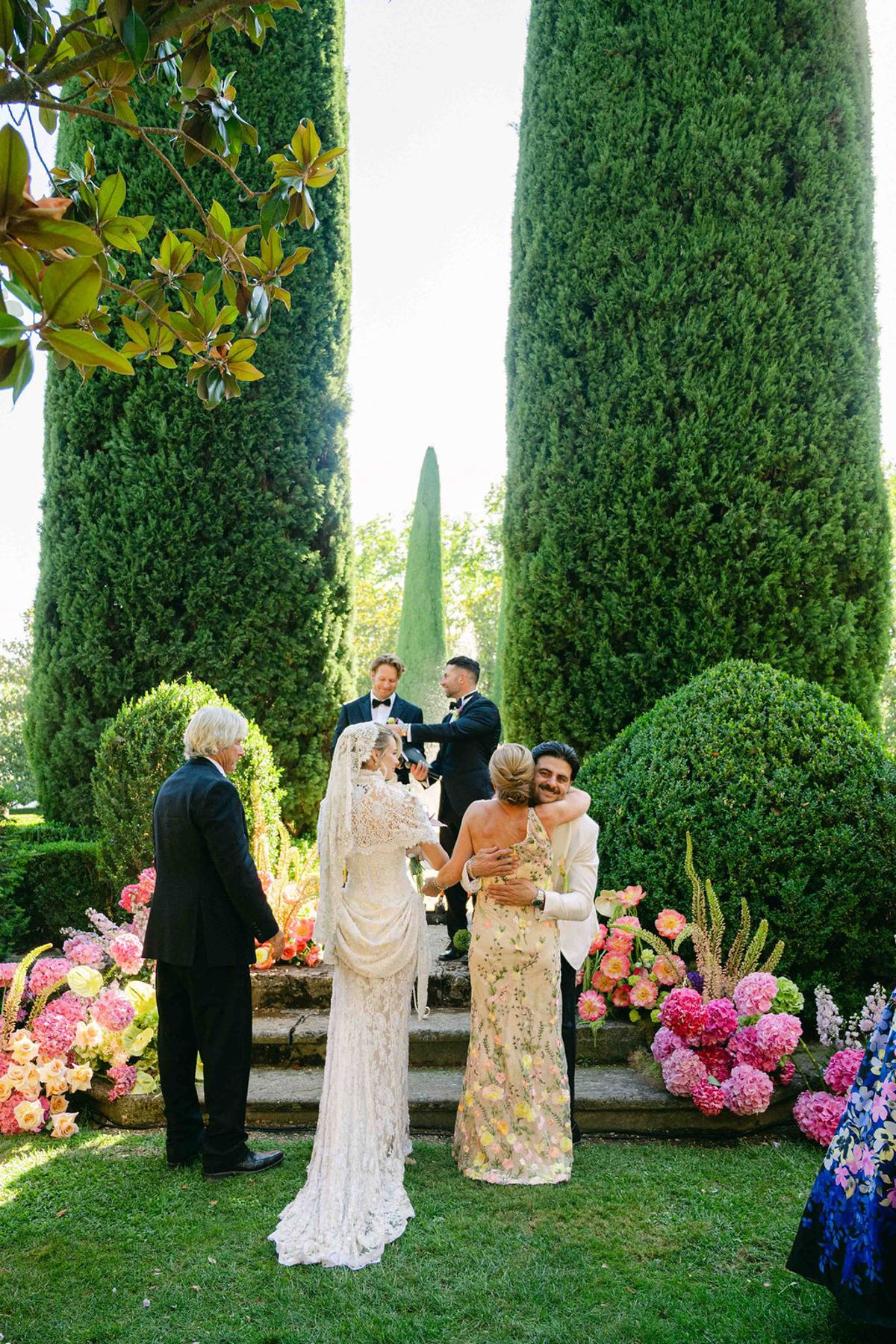 The bride and groom stand on flower-lined steps embracing a woman in a gold dress, with an older man beside them.