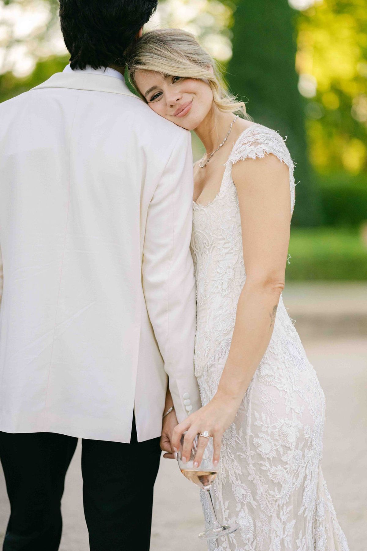 The bride in a lace dress rests her head against the groom's shoulder, smiling, while holding a glass in a soft green setting.