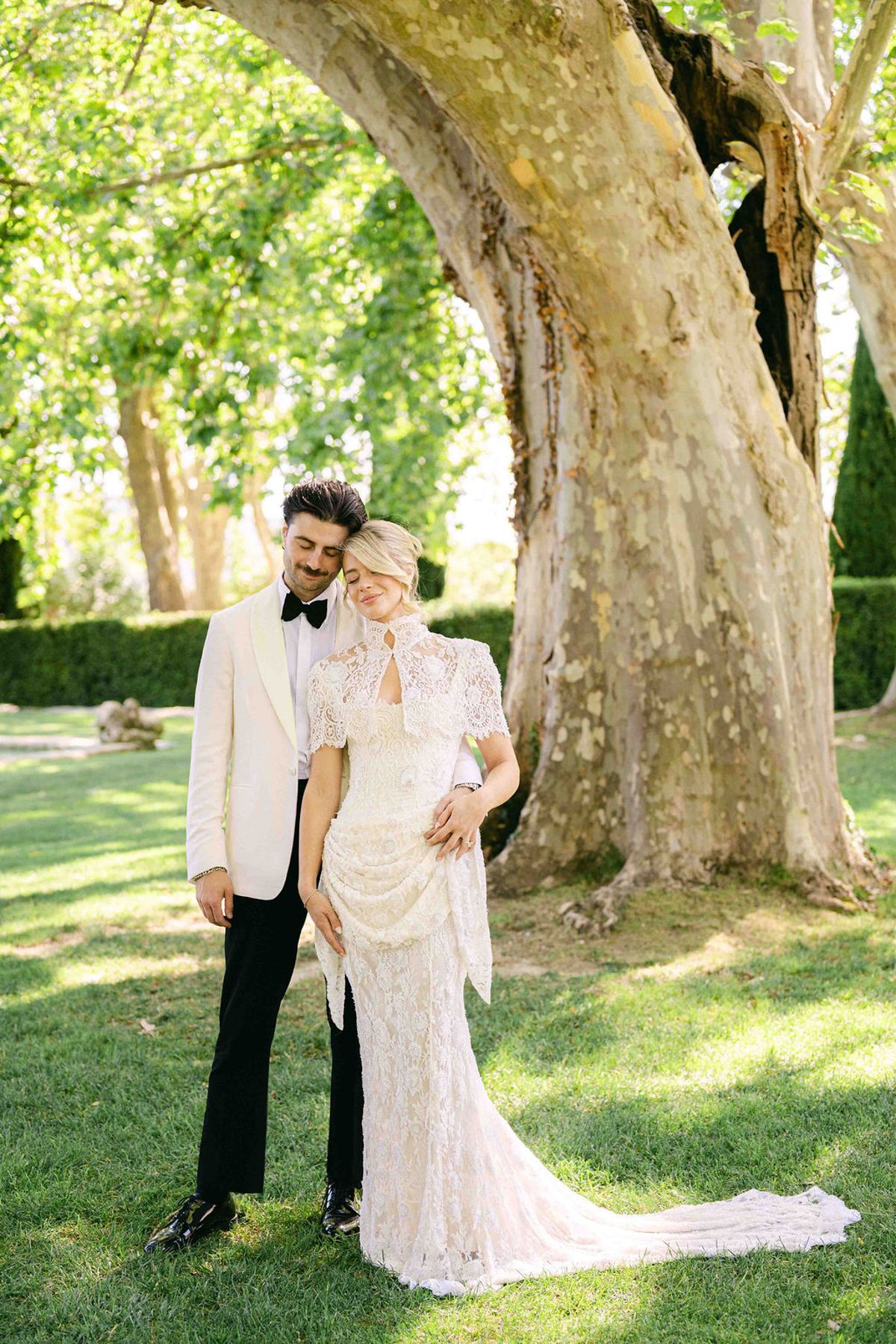 The groom in a white jacket kisses the bride's head as they stand on a lawn beside a large tree trunk.
