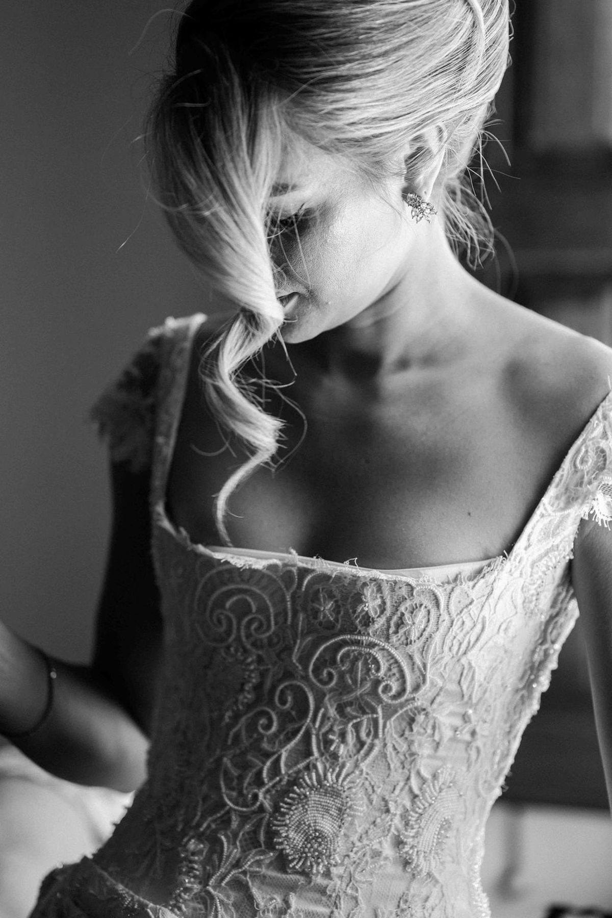 In black and white, the bride looks down with strands of hair across her face, wearing a lace-embroidered bodice.