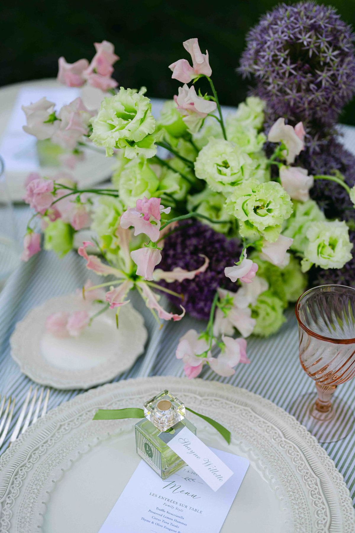A place setting with scalloped plates, a printed menu and a small green favour sits beside a bouquet of green, pink and purple flowers.