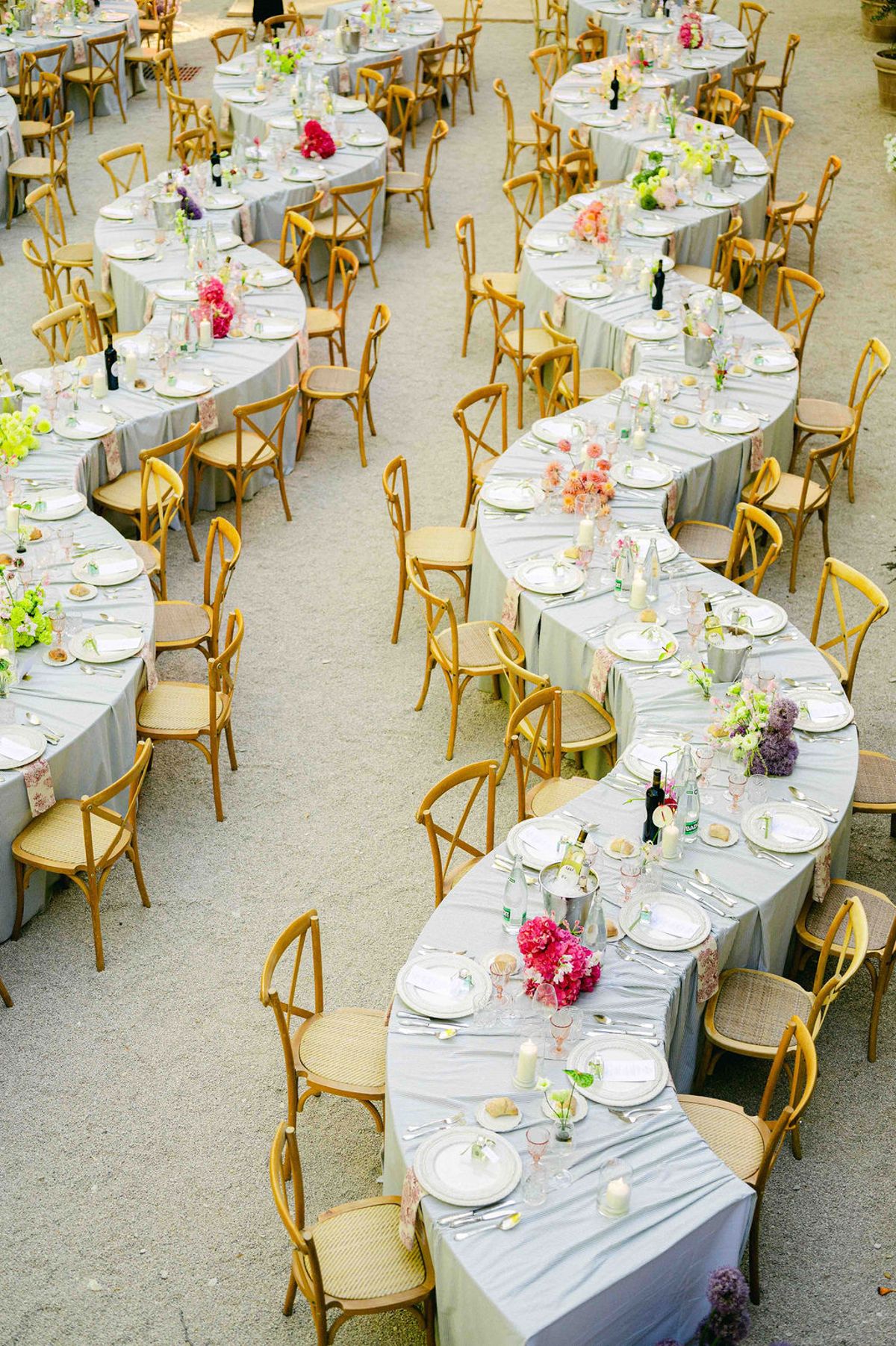 Seen from above, a long winding banquet table with grey linen, wooden chairs and small floral arrangements curves across a gravel courtyard.