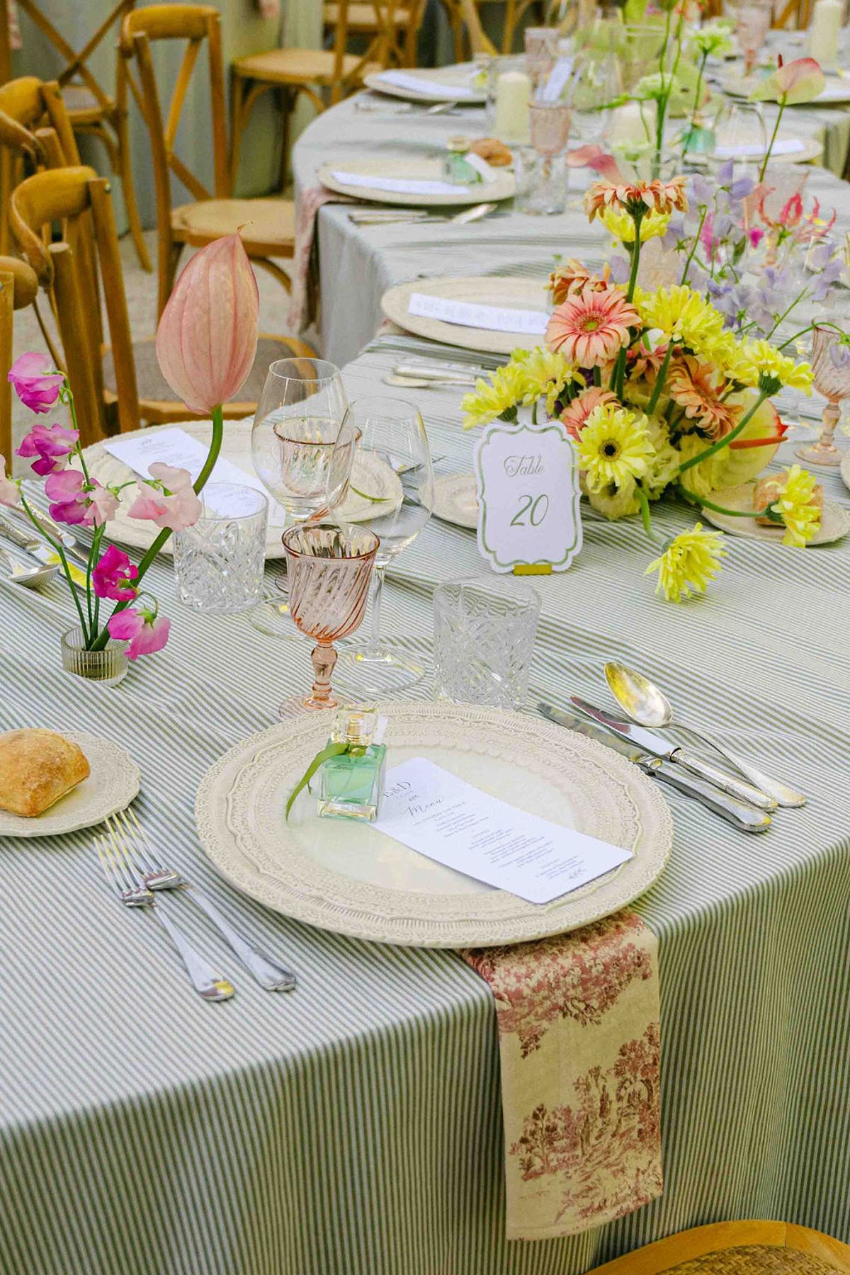 A round table with a striped cloth, pink glasses, a menu on a plate and a bouquet of yellow and pink flowers.