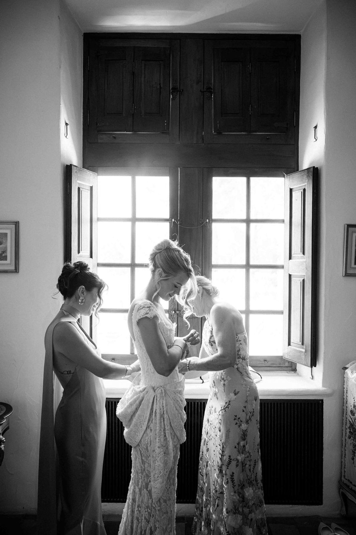 In black and white, two women help the bride with her dress in front of a large window with open shutters.