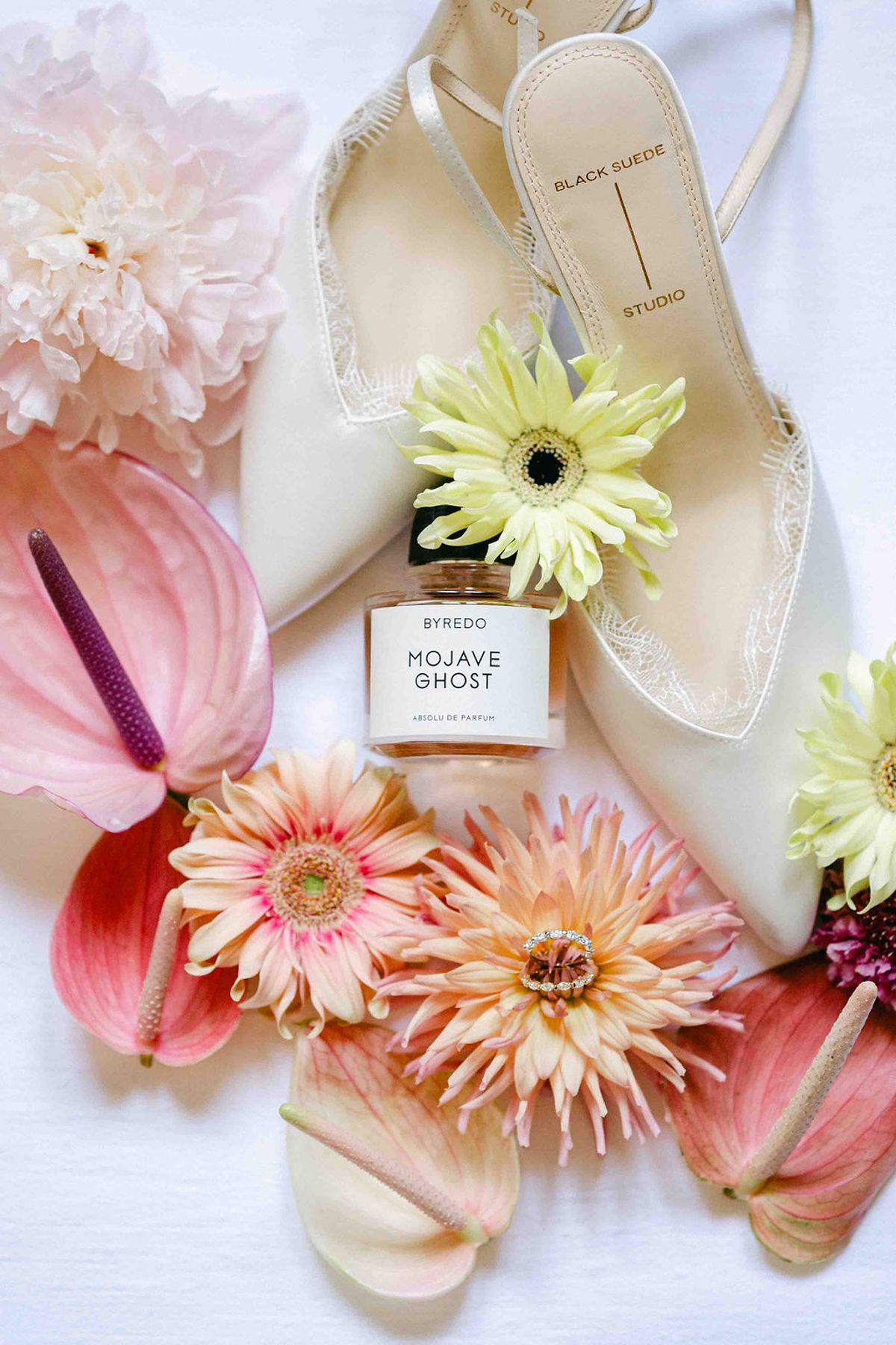 Cream bridal heels, a perfume bottle and pink and yellow gerberas and anthuriums are arranged on a white surface, seen from above.