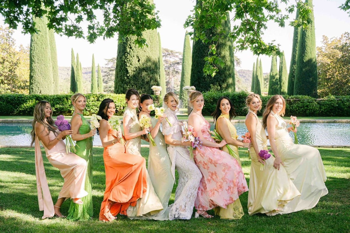 Ten women in green, orange, pink floral and yellow dresses stand in a line leaning together on a lawn beside a pool.