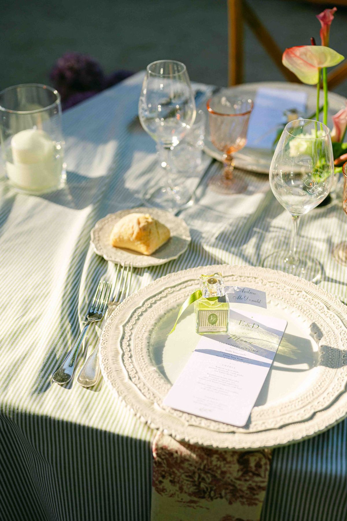 A scalloped plate with a menu card, bread roll and glasses sits on a striped tablecloth in patches of sunlight.