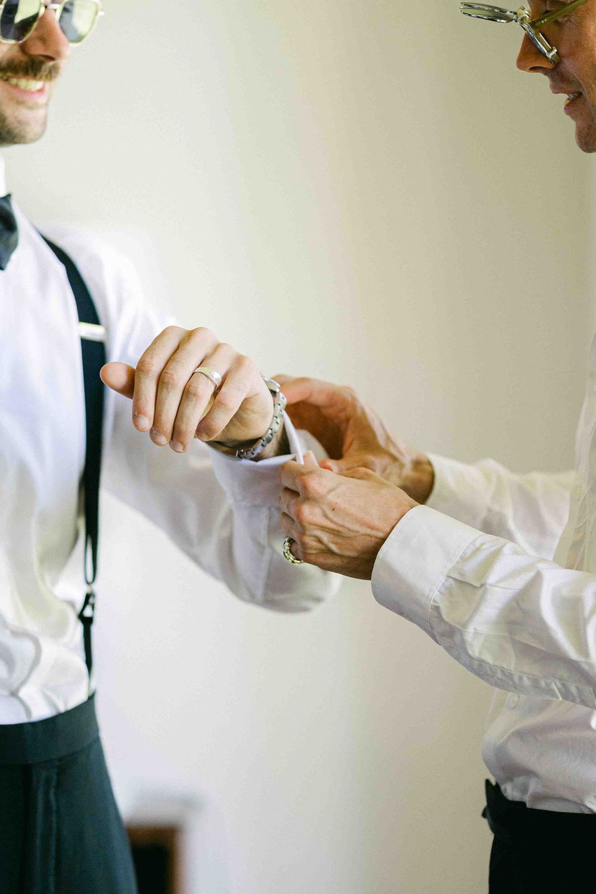 A man in a white shirt and braces has his cuff fastened by a second person, in a bright room.