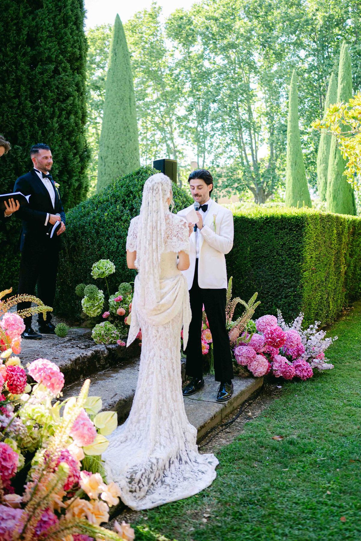 The bride in a long lace gown and veil faces the groom in a white jacket as an officiant stands nearby.