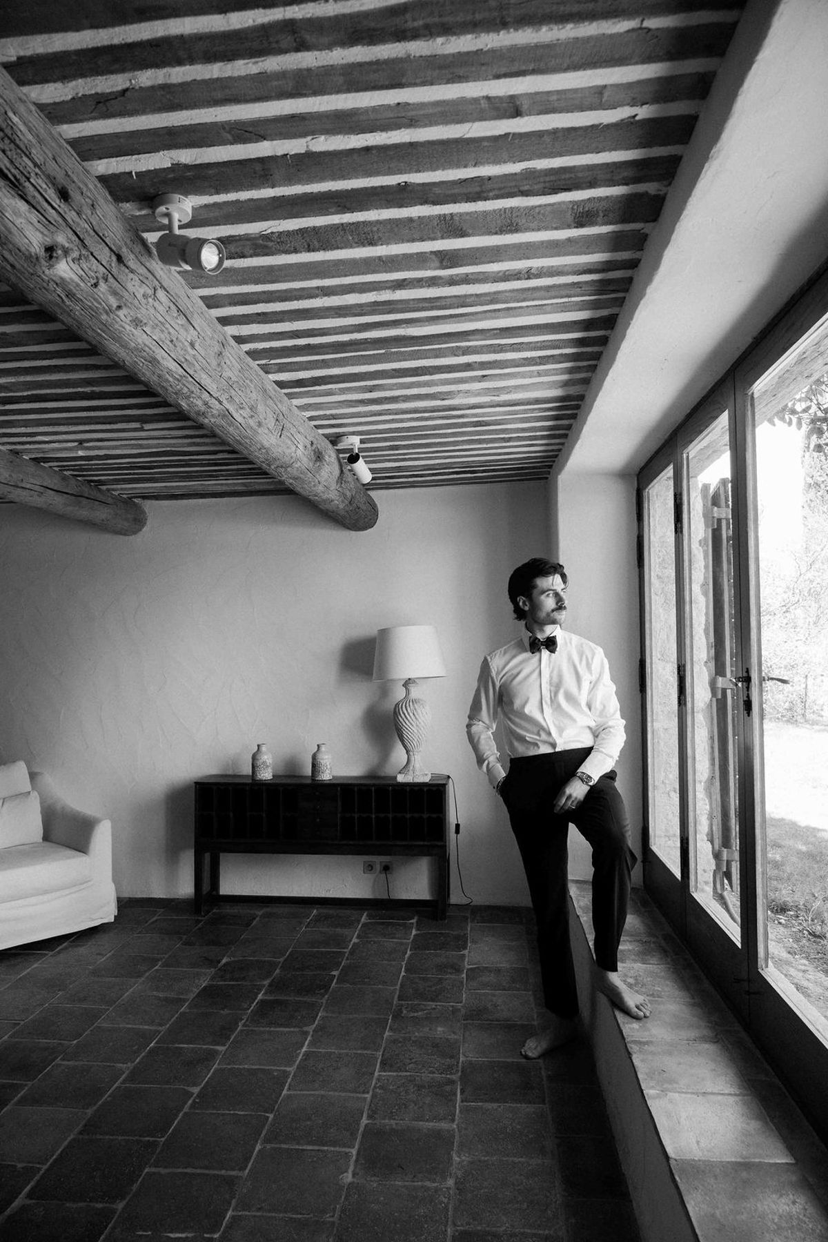 A man in a white shirt and bow tie stands beside a large window in a room with a wooden beamed ceiling.