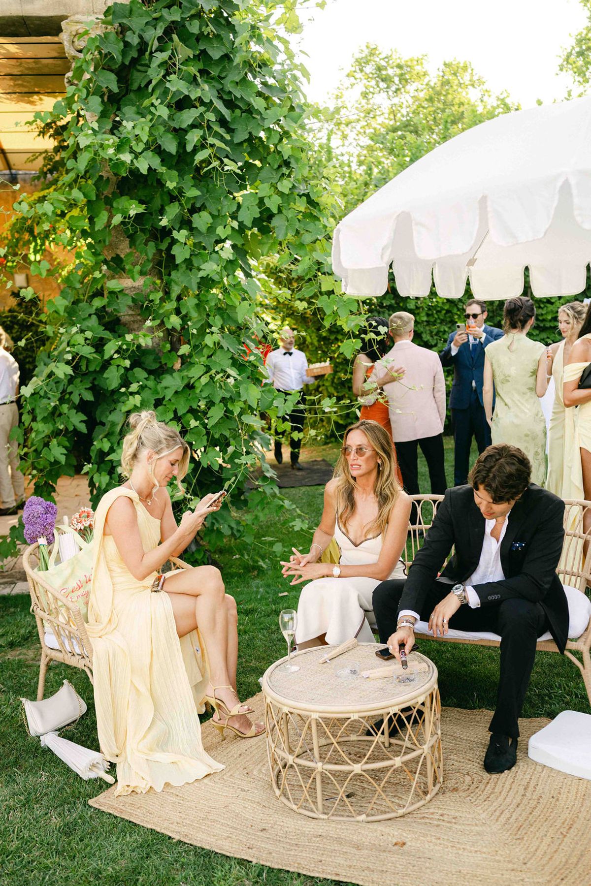 Guests sit and stand around a rattan table on a rug in a garden beneath a white fringed parasol.