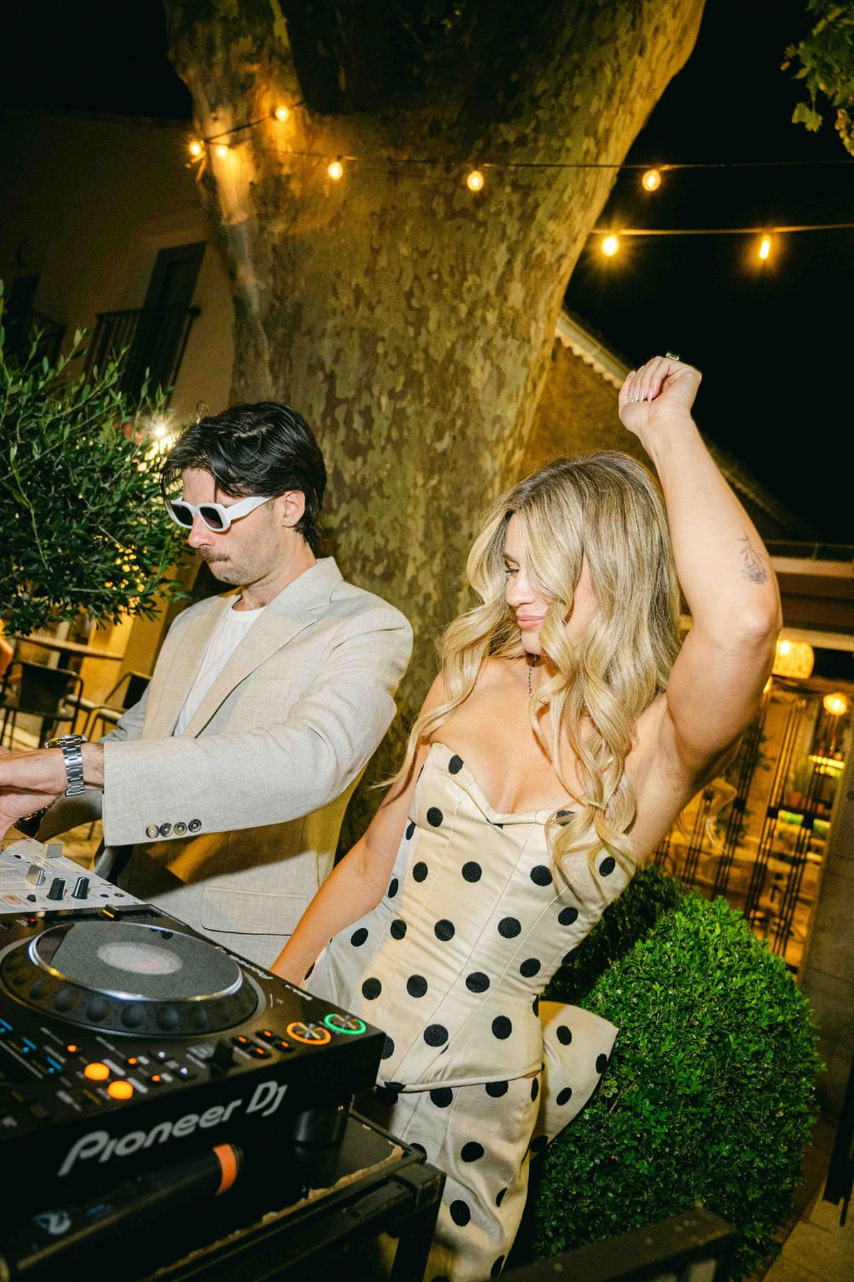 At night under string lights, a man in sunglasses works a DJ deck while a woman in a polka dot dress dances with her arm raised.