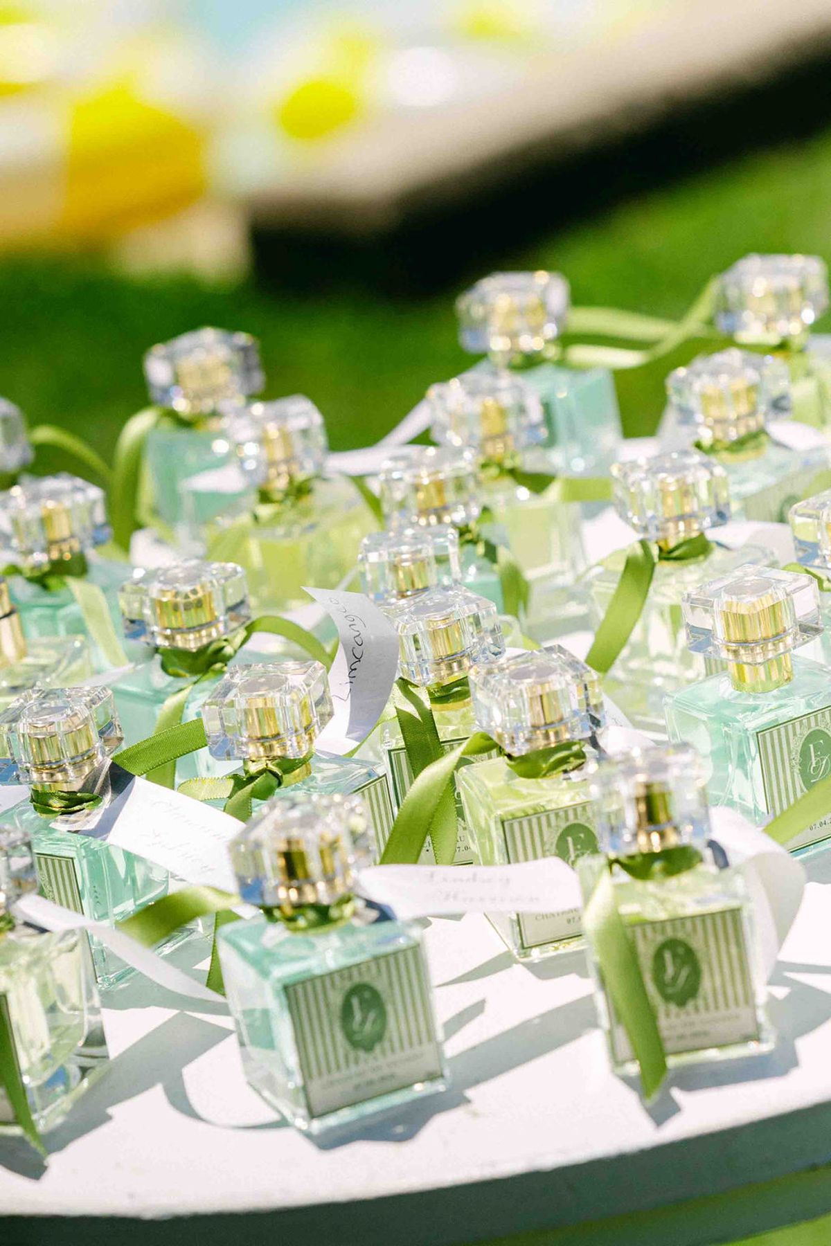 Rows of small glass perfume bottles tied with green ribbon sit on a white tray set on grass.