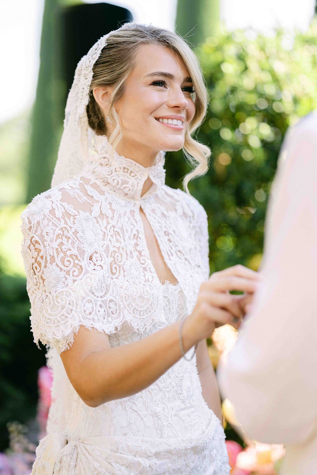 The bride, wearing a lace jacket and a veil, smiles while holding hands with a person whose back is partly visible in the foreground.