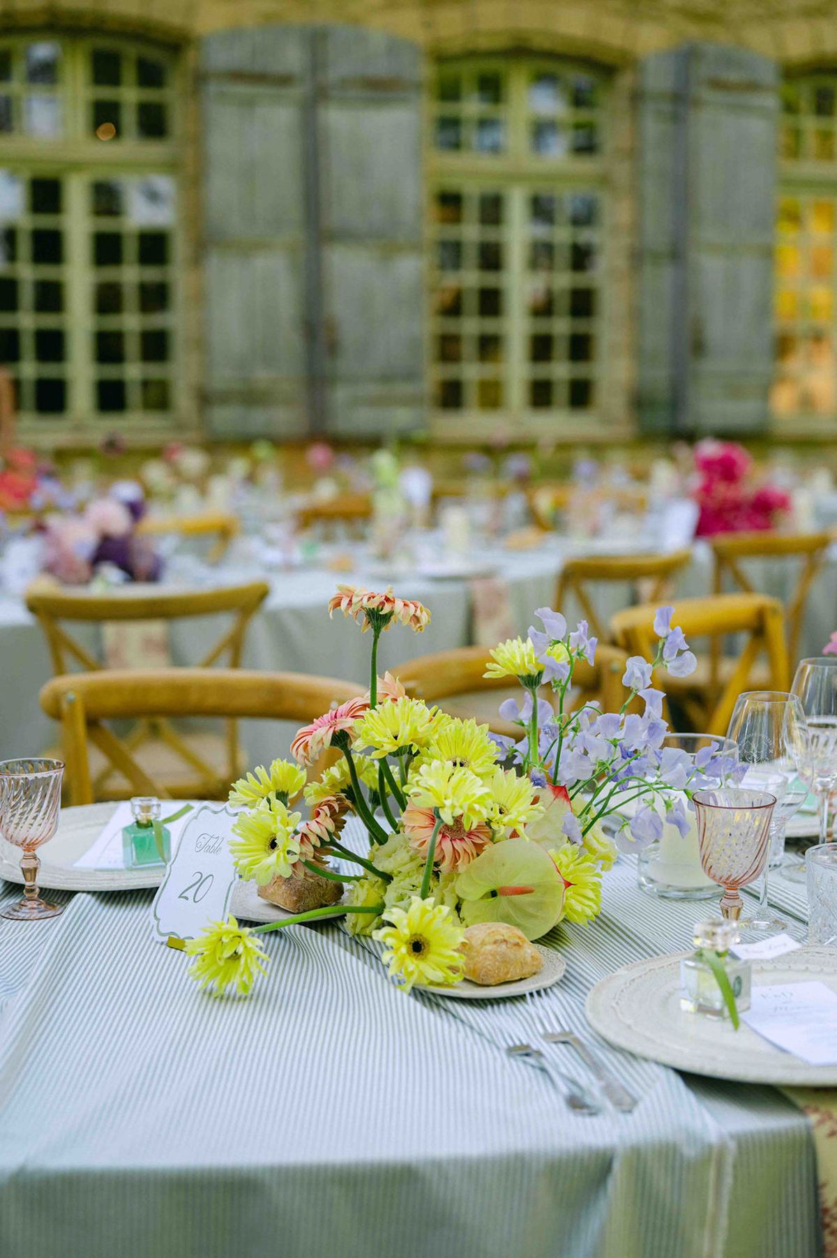 Tables with pale blue cloths, yellow flower arrangements and pink glasses stand in front of a chateau with shuttered windows.