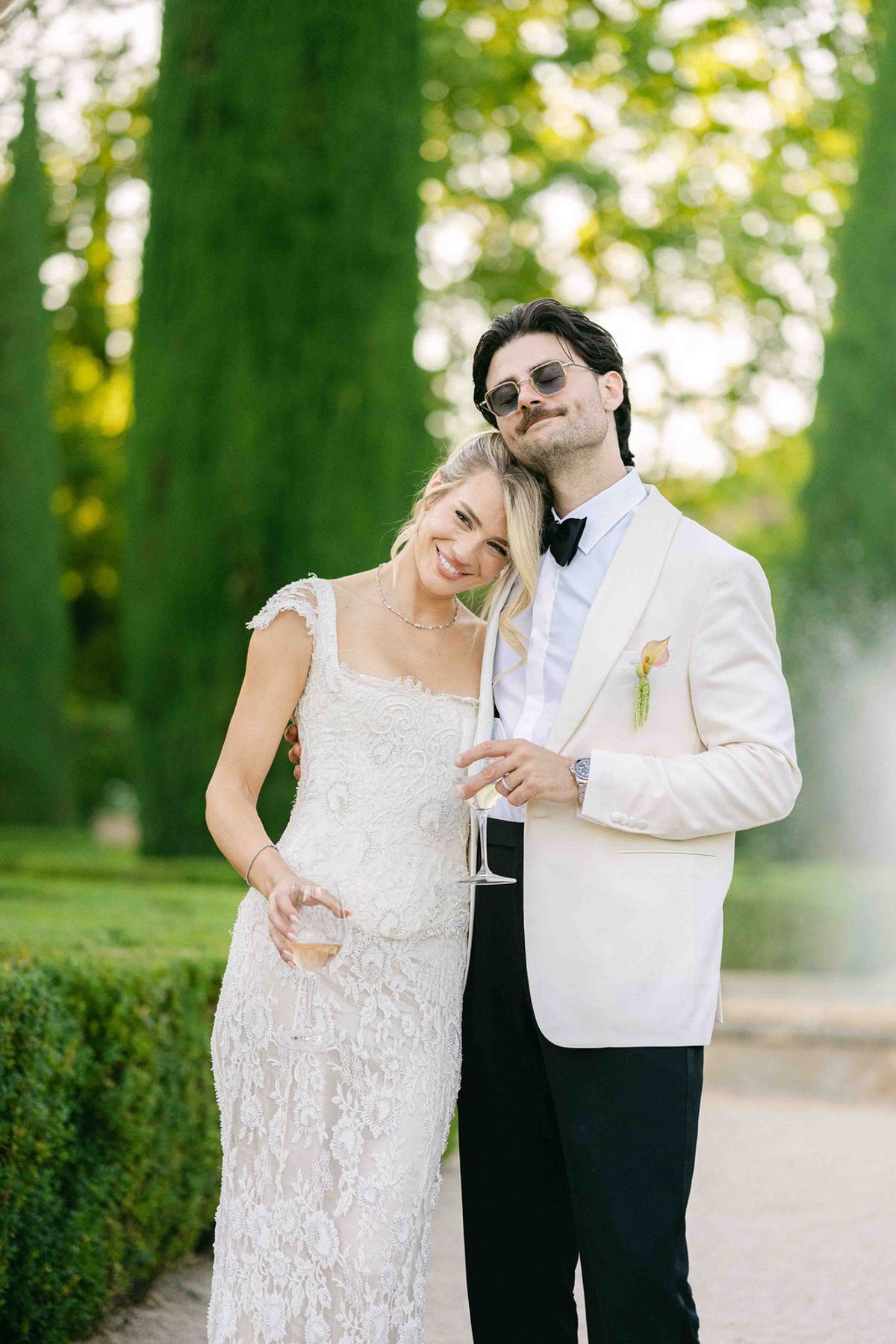 The couple stand close together smiling in a sunlit garden, the groom in a white jacket and sunglasses, both holding glasses.