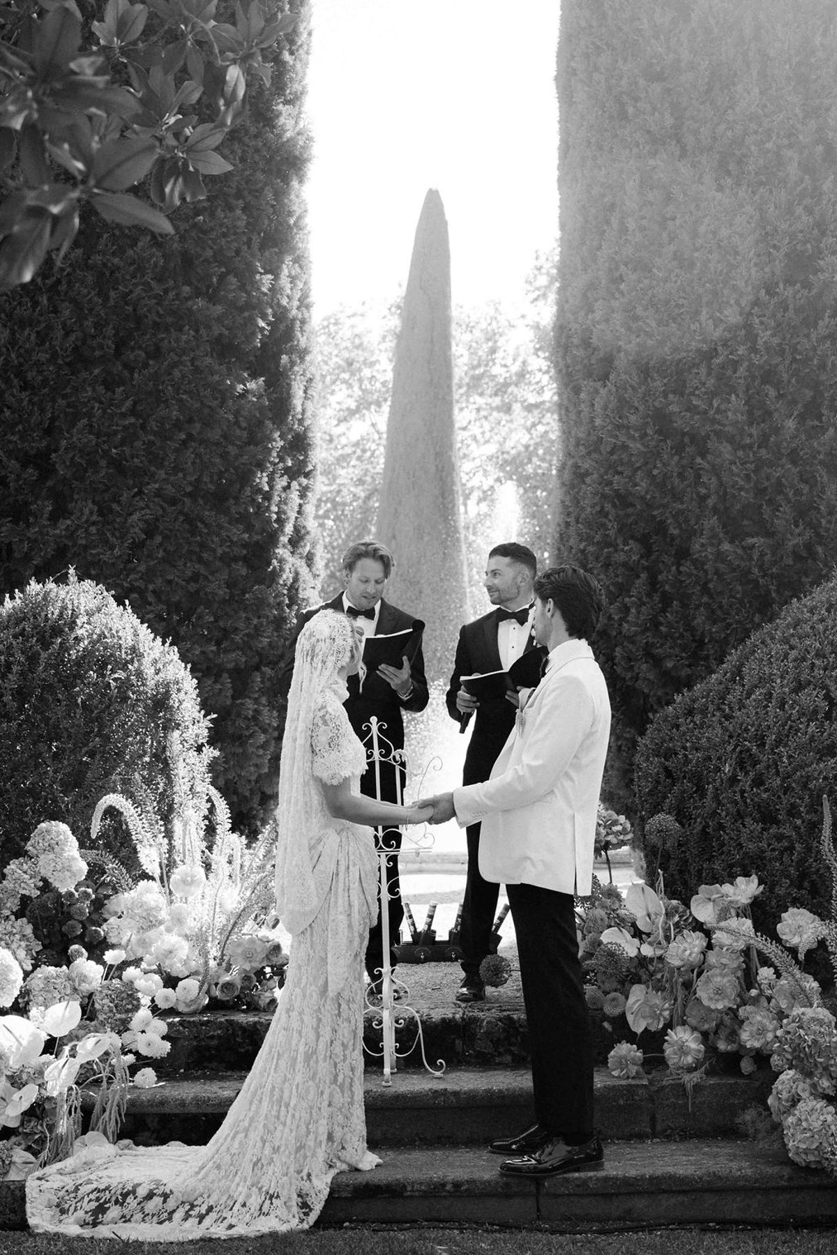 In black and white, the bride and groom hold hands facing an officiant between tall cypress trees and large floral arrangements.